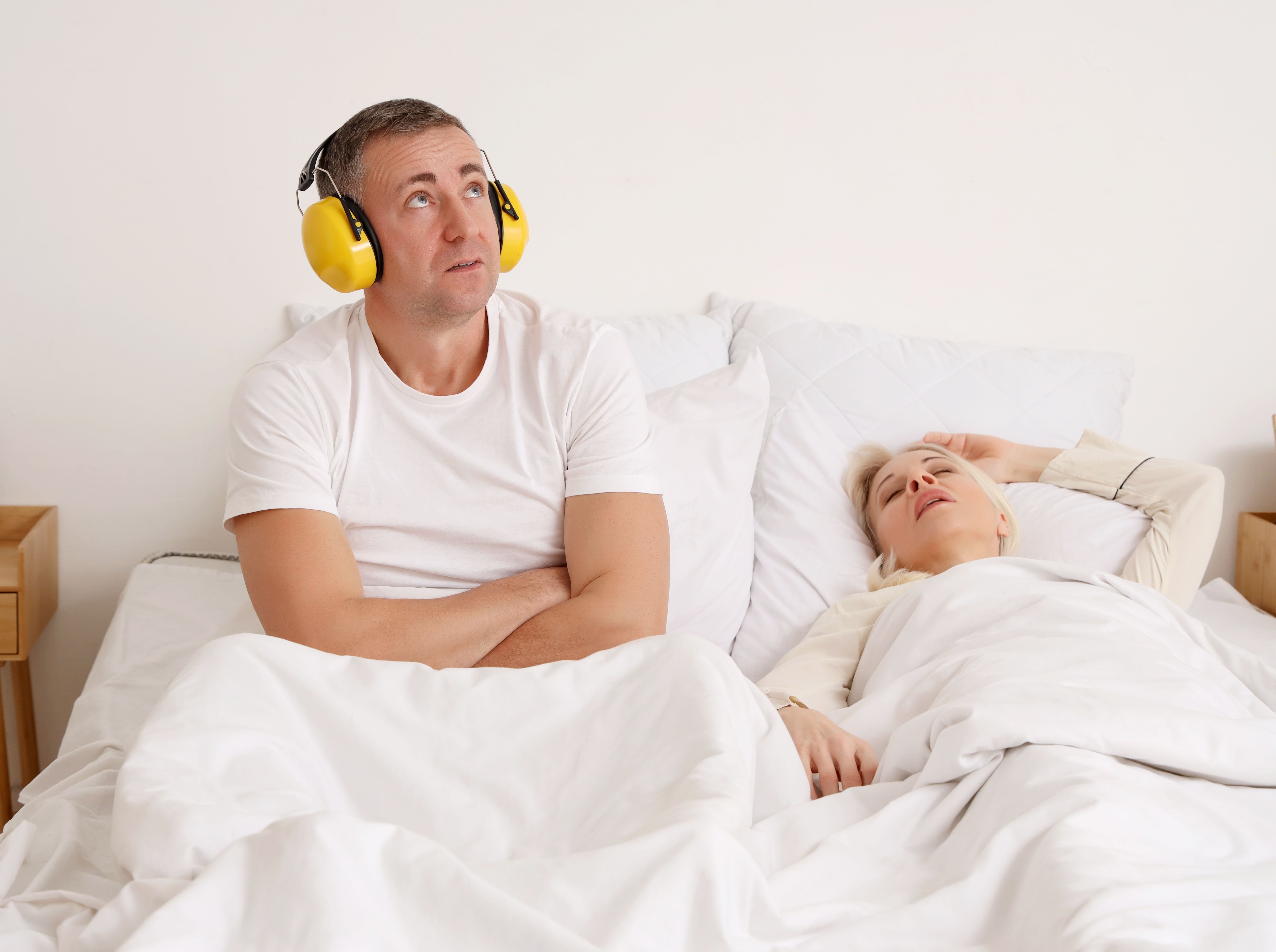Mature man with hearing protectors cannot sleep because of his wife's snoring in bedroom