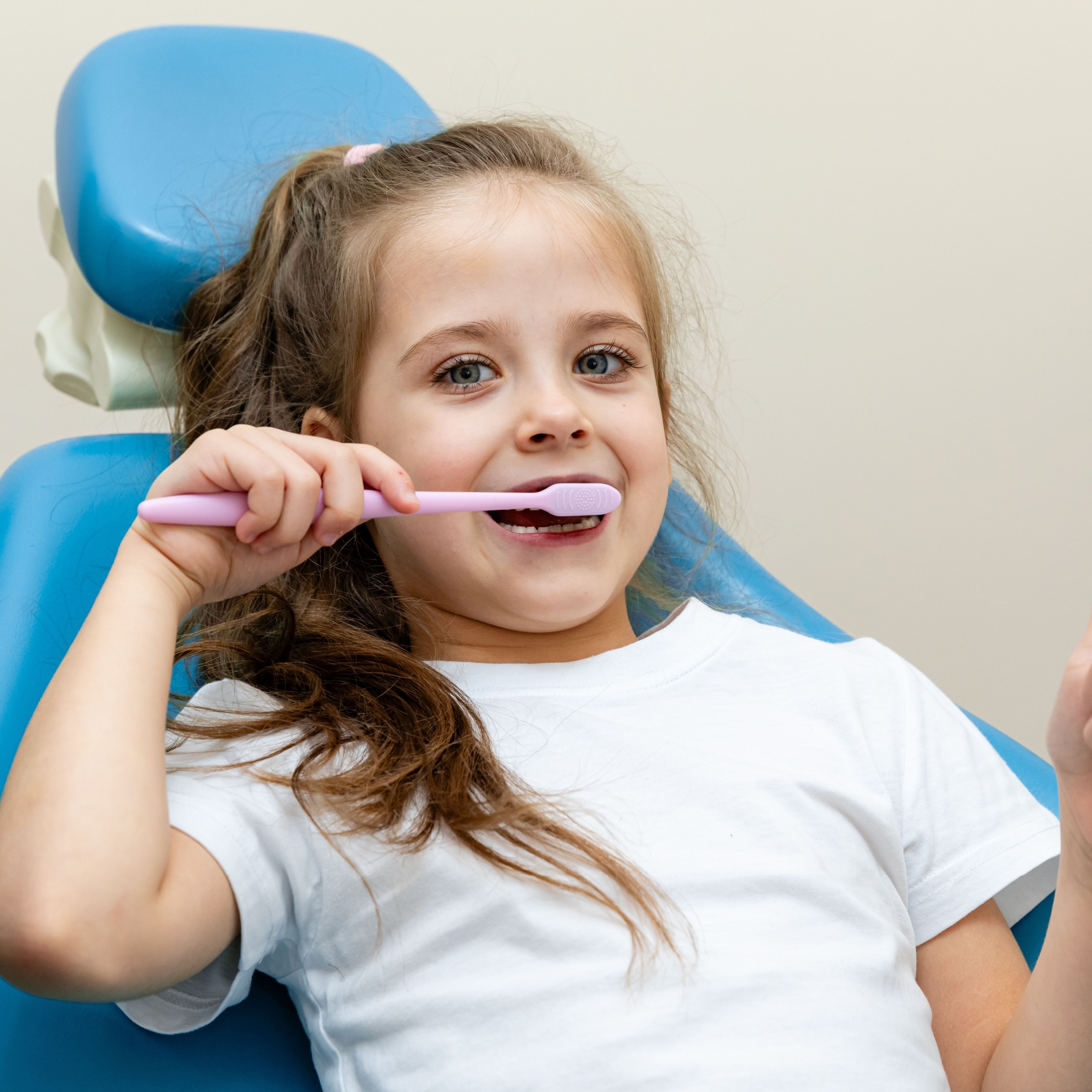 Cute little girl sitting in dentist's chair, brushing her teeth and showing thumbs up.