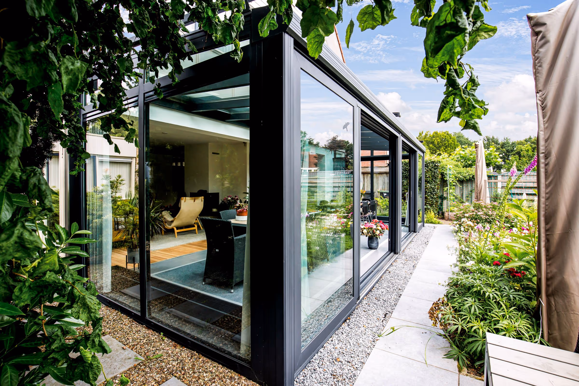 Moderne aluminium serre met glazen wanden, geplaatst naast een woning in een groene tuin