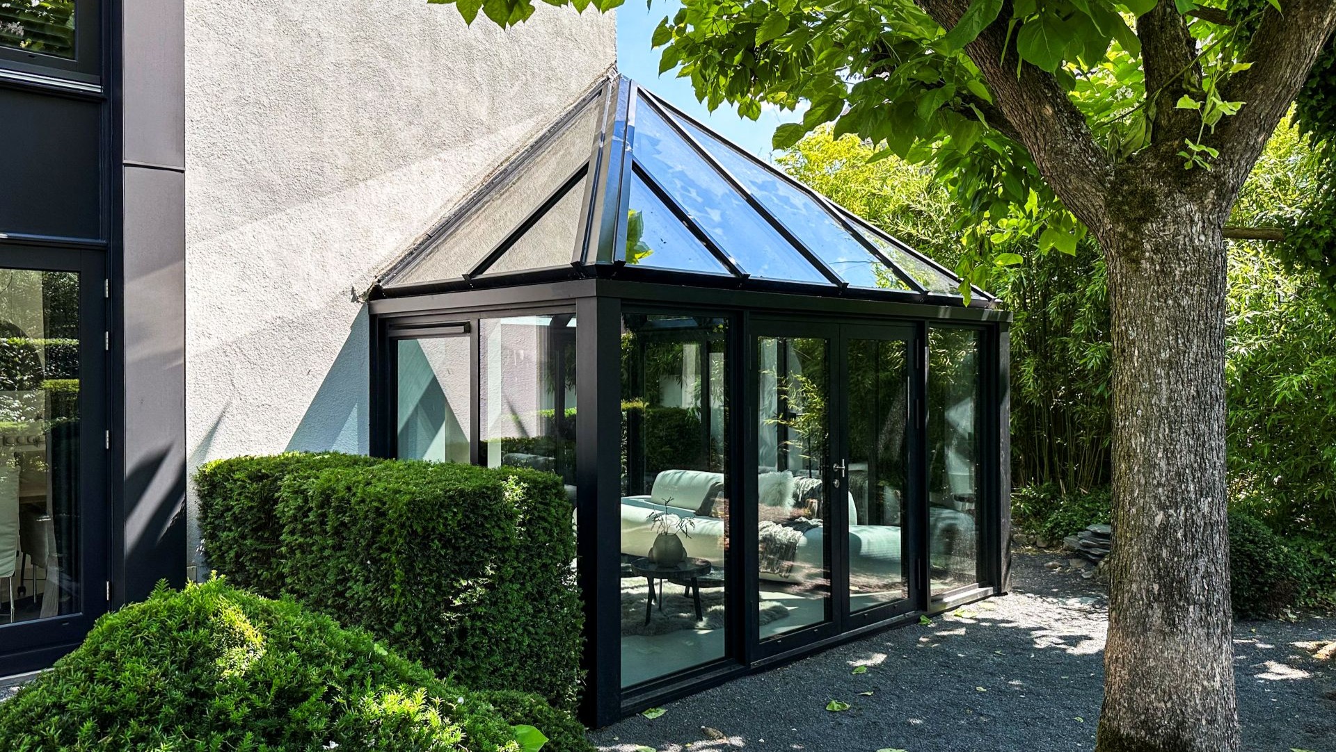 Moderne zwarte aluminium serre met glazen dak en schuifpuien, geplaatst naast een woning in een groene tuin