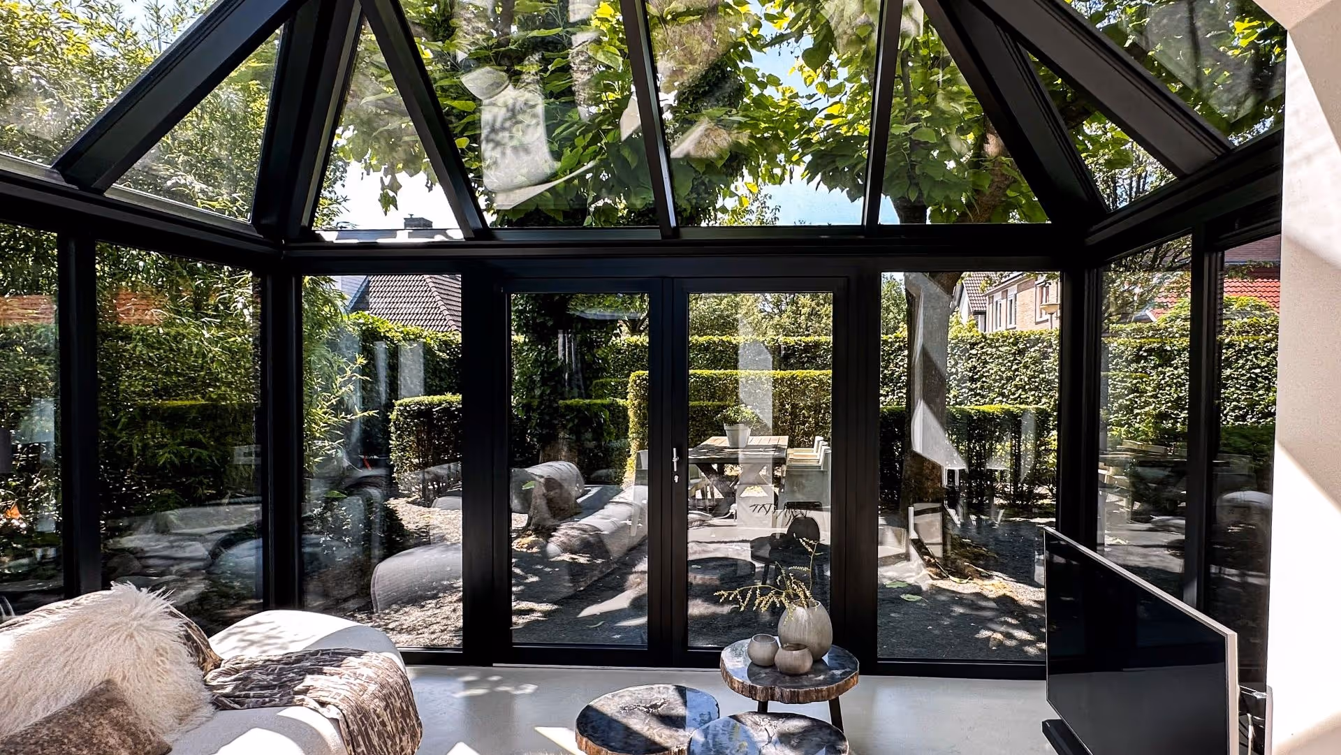 Uitzicht vanuit een serre met glazen wanden op een tuin met eettafel en groene hagen