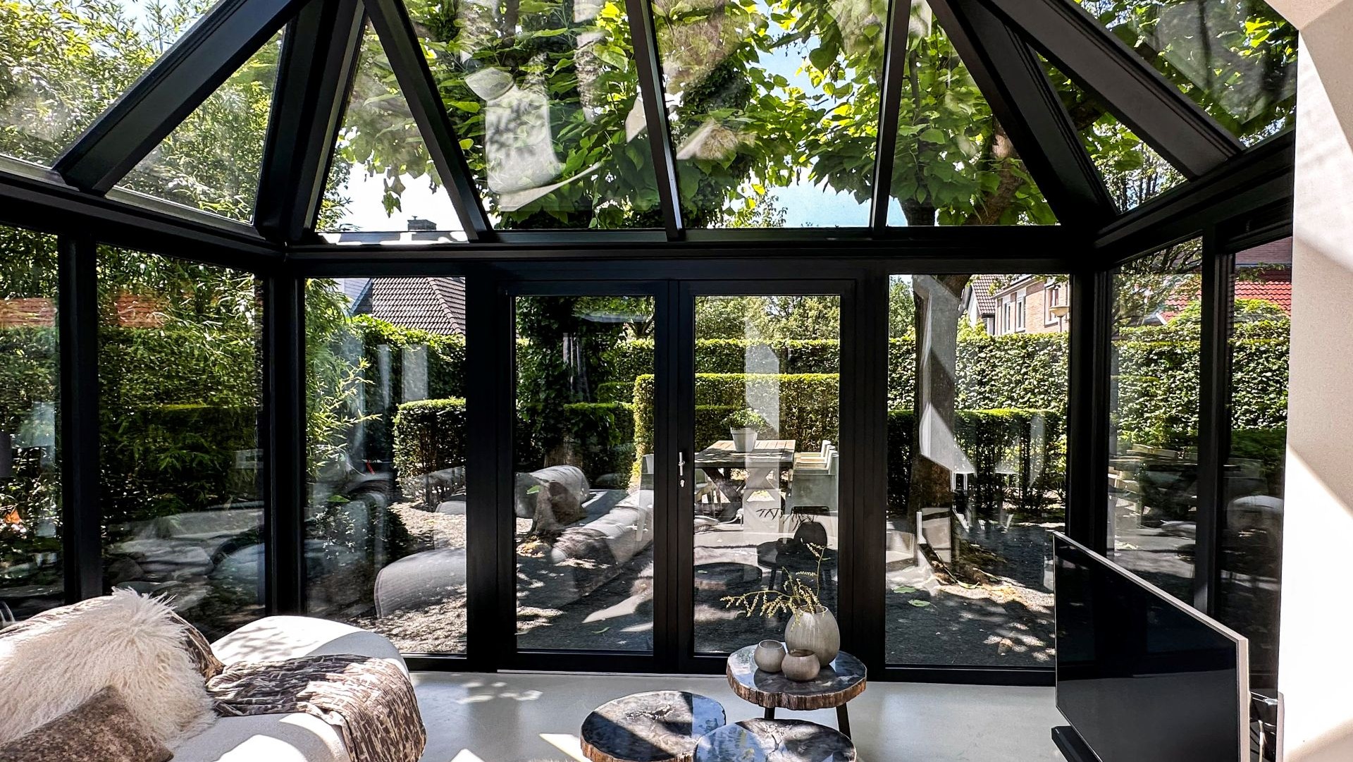 Uitzicht vanuit een serre met glazen wanden op een tuin met eettafel en groene hagen