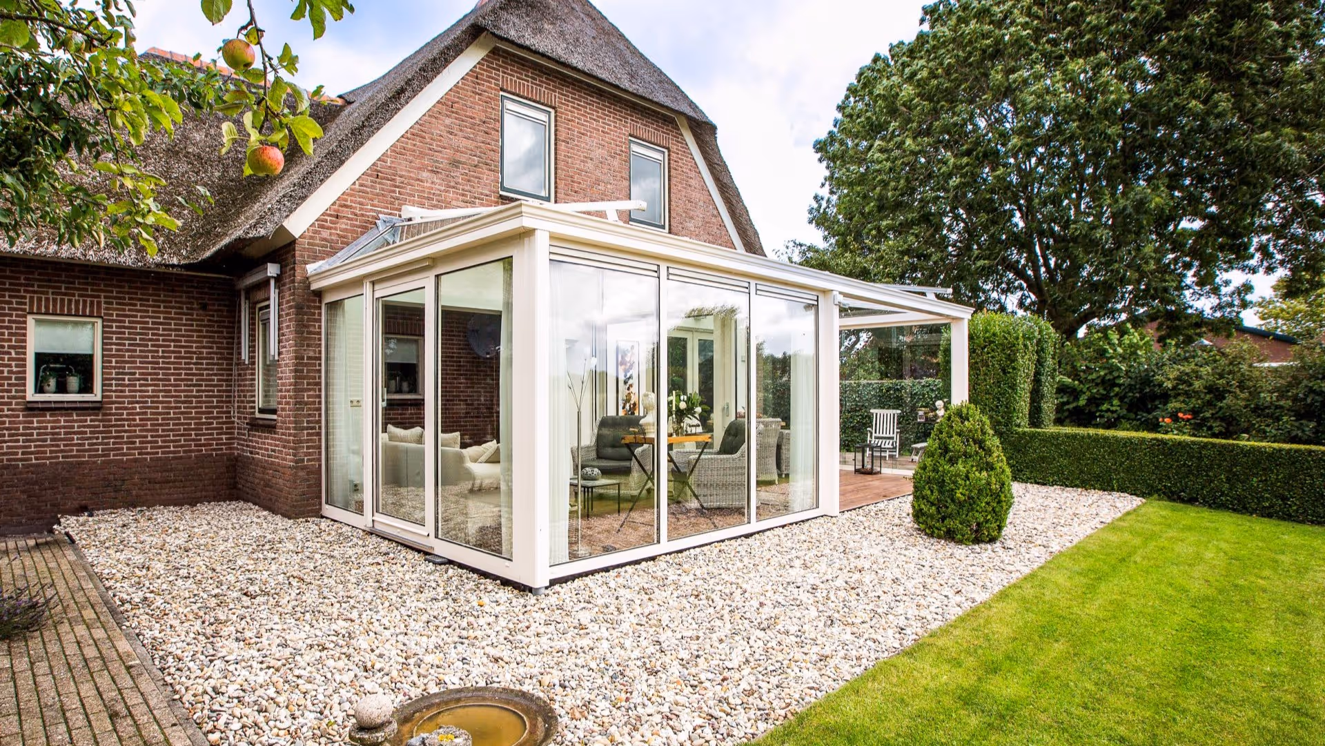 Ruime serre met glazen wanden en overkapping, geplaatst in een groene tuin bij een vrijstaande woning