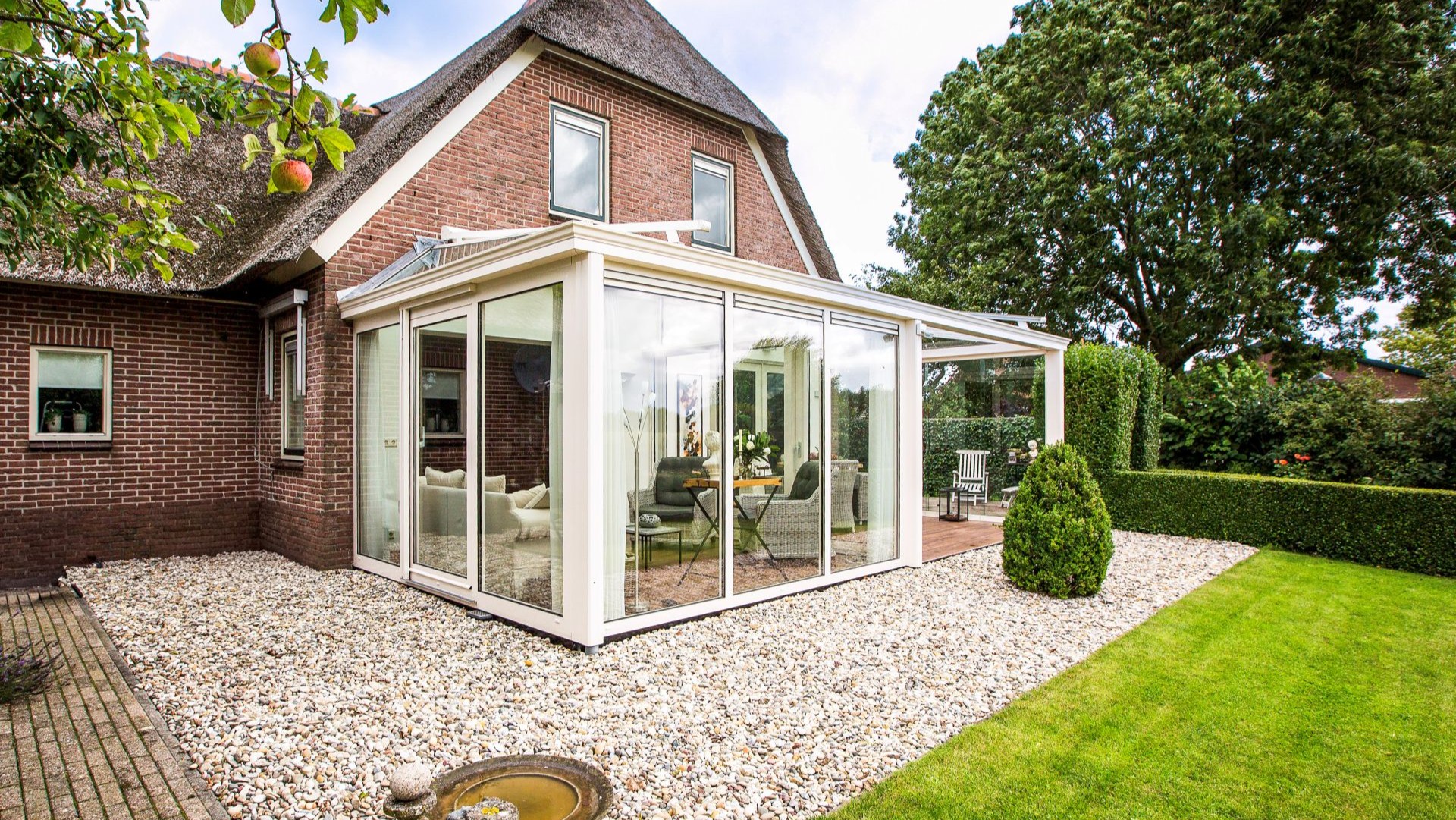 Ruime serre met glazen wanden en overkapping, geplaatst in een groene tuin bij een vrijstaande woning