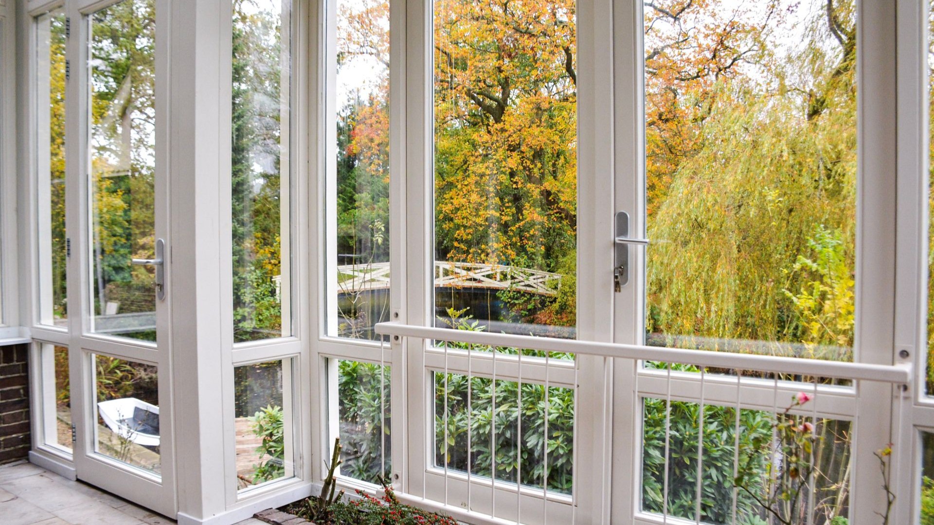 Uitzicht vanuit serre met grote ramen op tuin, water en brug in herfstkleuren