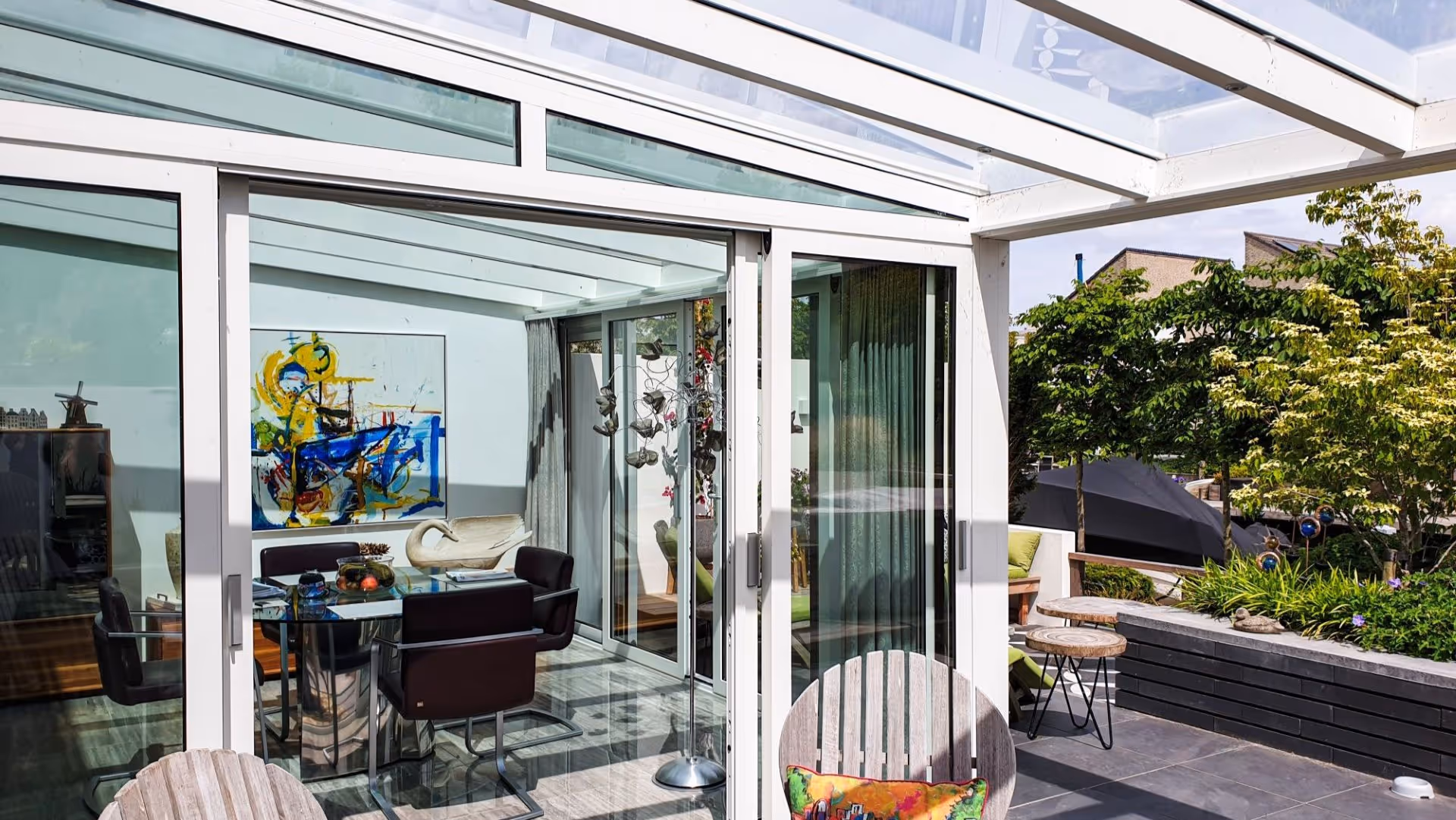Overdekt terras met glazen dakconstructie, loungeset en buitenkeuken, gelegen aan het water met uitzicht op woningen en tuin.