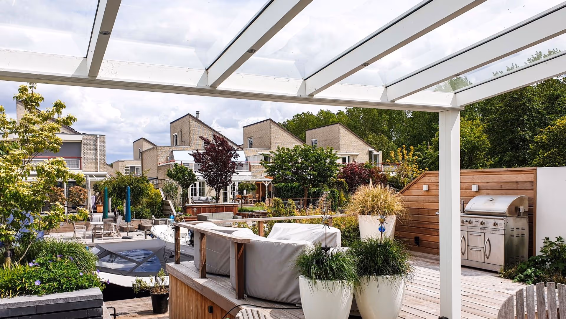 Luxe tuinkamer met glazen dak en open schuifpuien, met zicht op terras met buitenkeuken en groene beplanting.