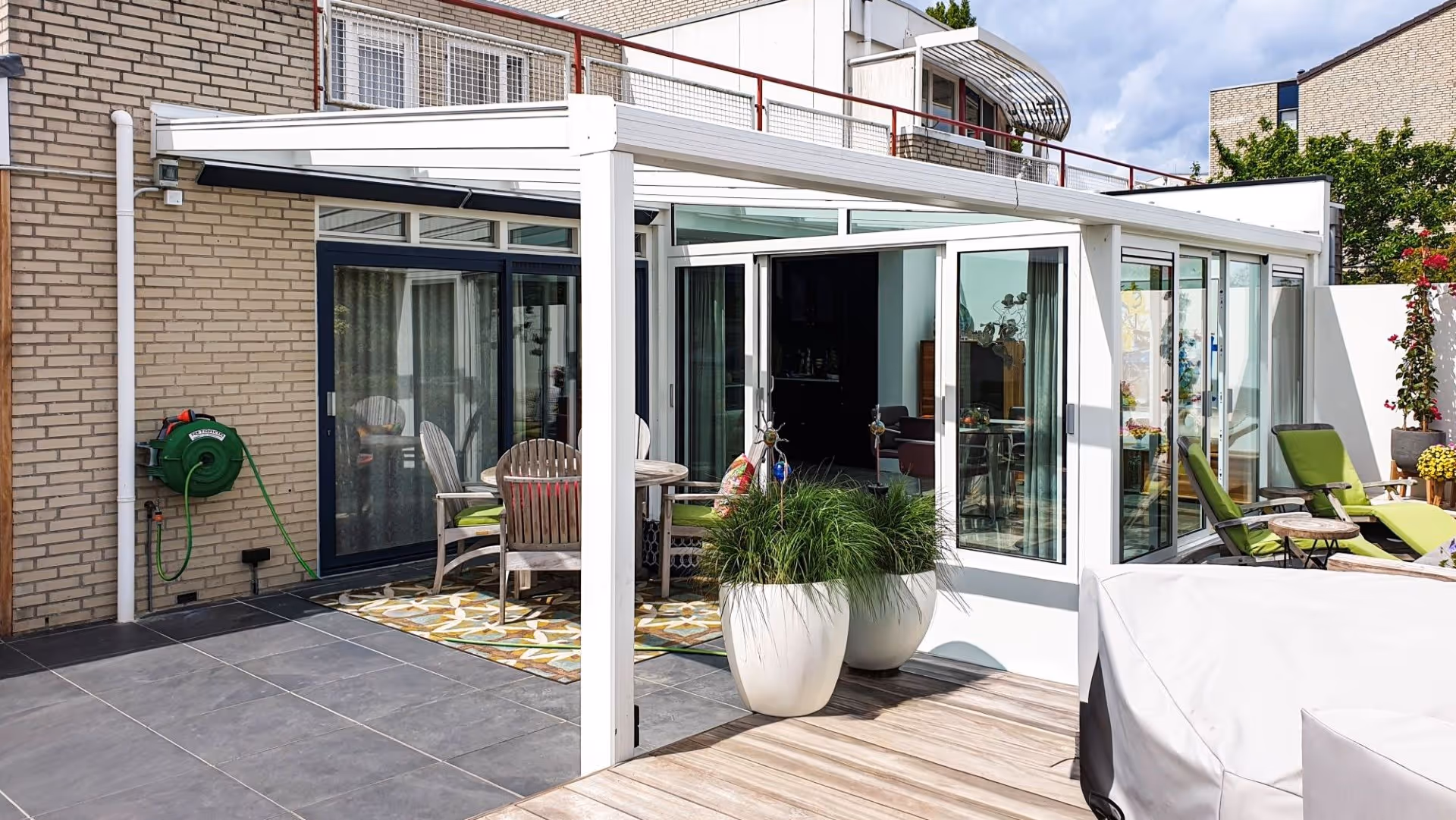 Terras met ligstoelen en groen aangelegde tuin, verbonden met moderne tuinkamer met glazen schuifpuien.