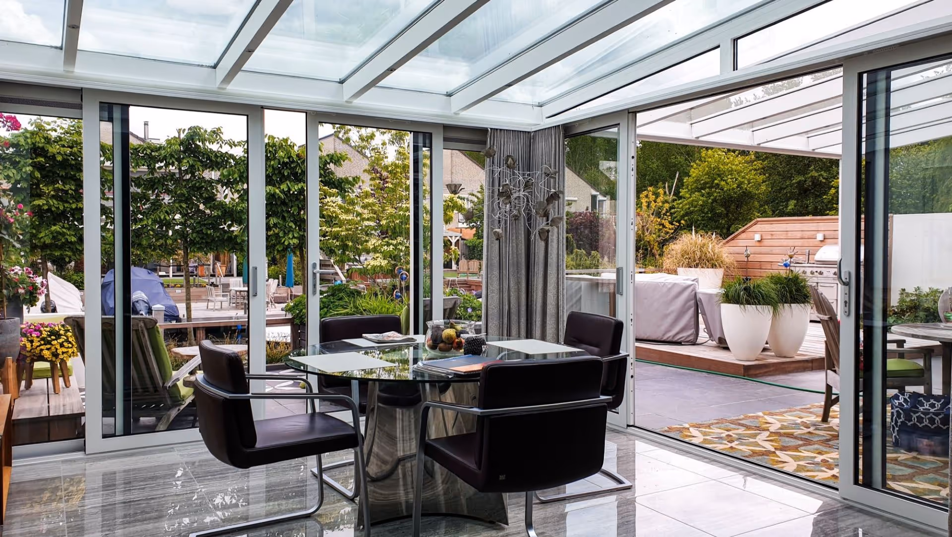 Moderne serre met glazen overkapping en schuifdeuren, ingericht met eettafel en stoelen, verbonden met luxe tuin en buitenruimte.