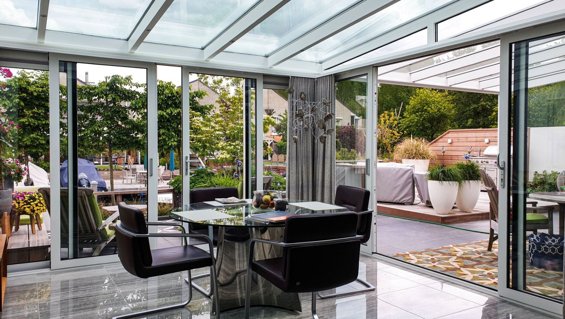 Moderne serre met glazen overkapping en schuifdeuren, ingericht met eettafel en stoelen, verbonden met luxe tuin en buitenruimte.