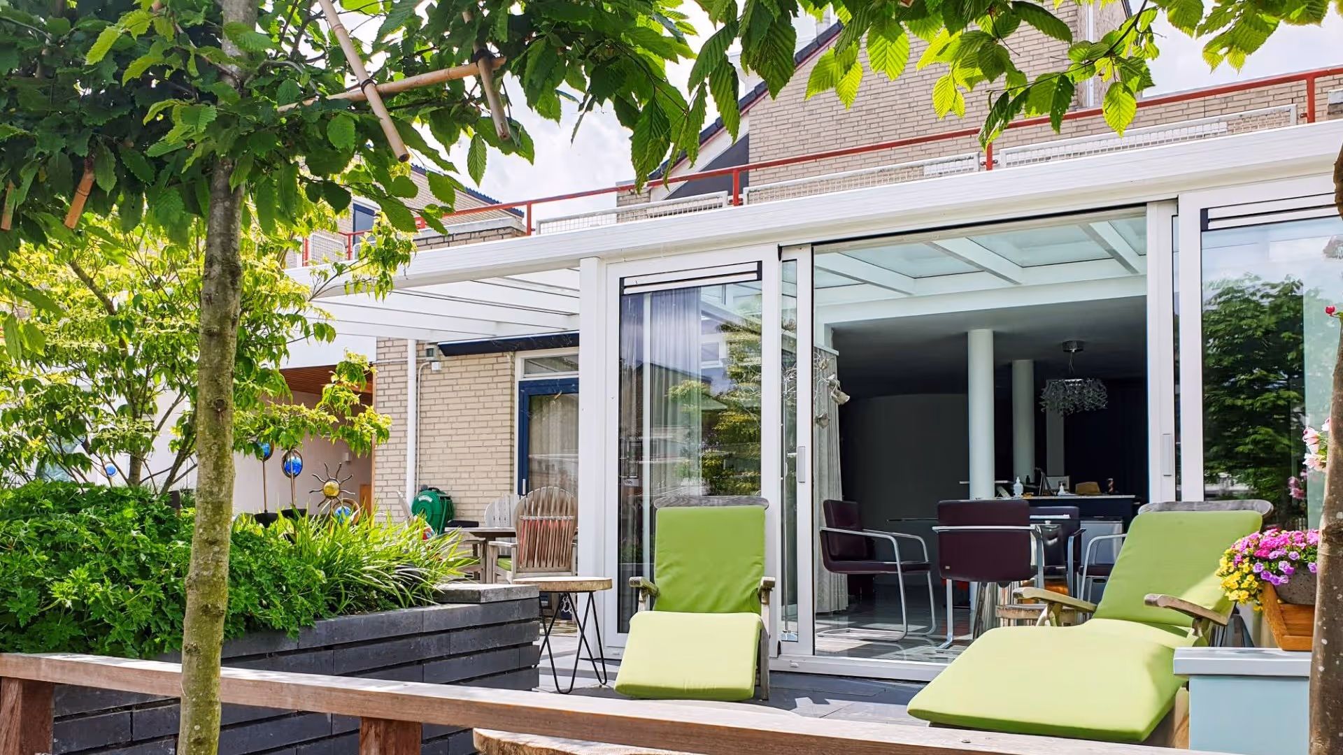 Tuinkamer met glazen wanden en dak, voorzien van eethoek en uitzicht op groene tuin met terras aan het water.