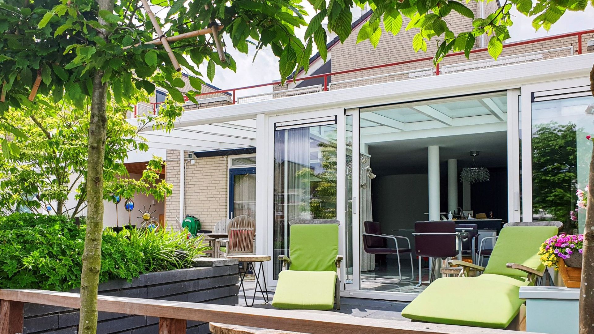 Tuinkamer met glazen wanden en dak, voorzien van eethoek en uitzicht op groene tuin met terras aan het water.