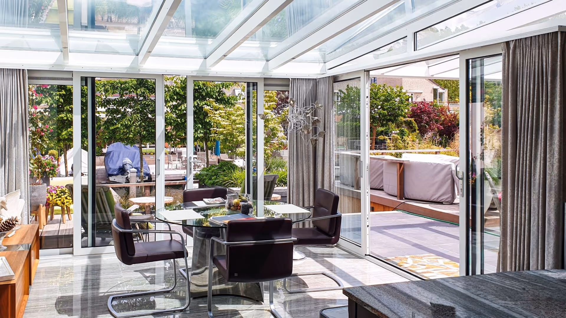 Ruime tuinkamer met glazen dak en aluminium schuifwanden, voorzien van moderne eethoek en uitzicht op sfeervolle tuin en terras.