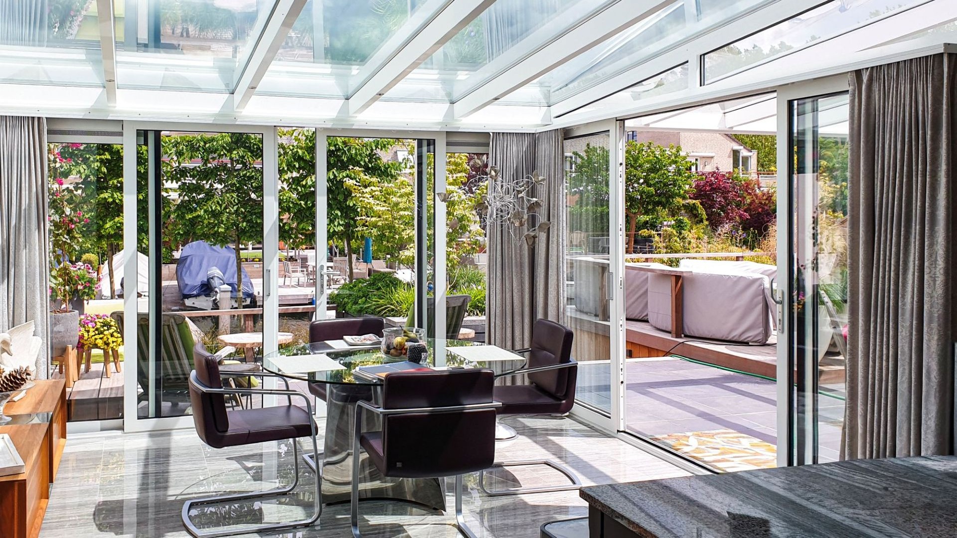 Ruime tuinkamer met glazen dak en aluminium schuifwanden, voorzien van moderne eethoek en uitzicht op sfeervolle tuin en terras.