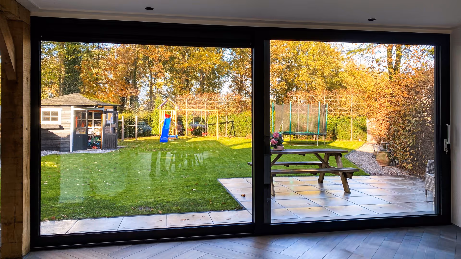 Uitzicht door een minimalistische schuifpui op een ruime tuin met terras, grasveld en houten schuur