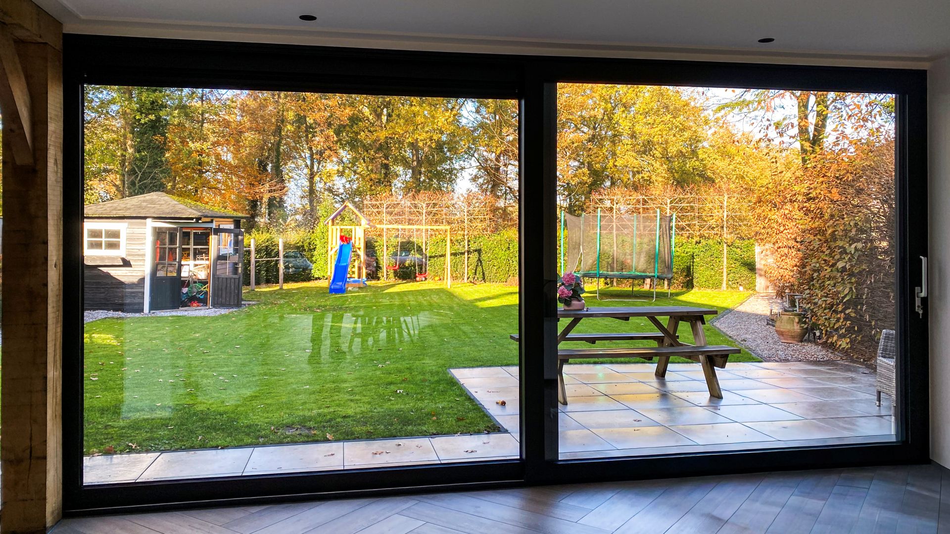 Uitzicht door een minimalistische schuifpui op een ruime tuin met terras, grasveld en houten schuur