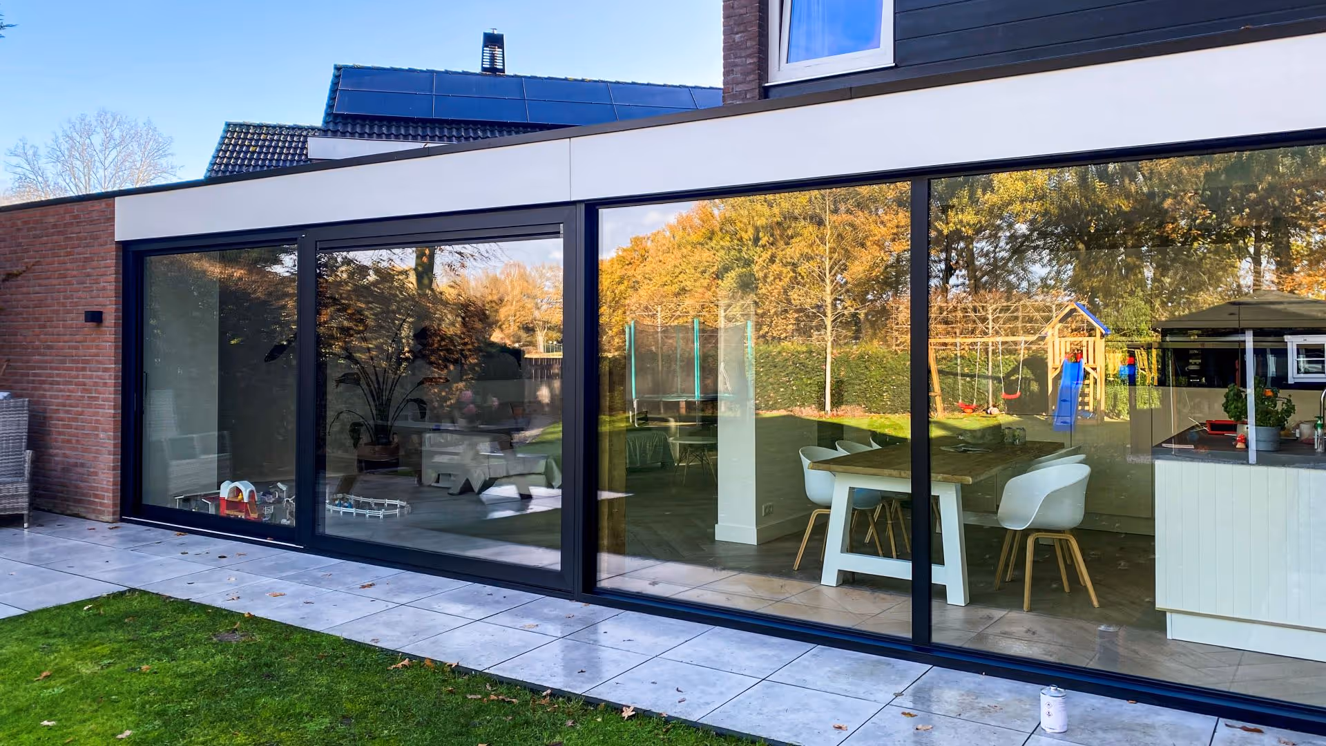 Brede aluminium schuifpui over de volledige gevel met uitzicht op een groene tuin en speeltoestellen