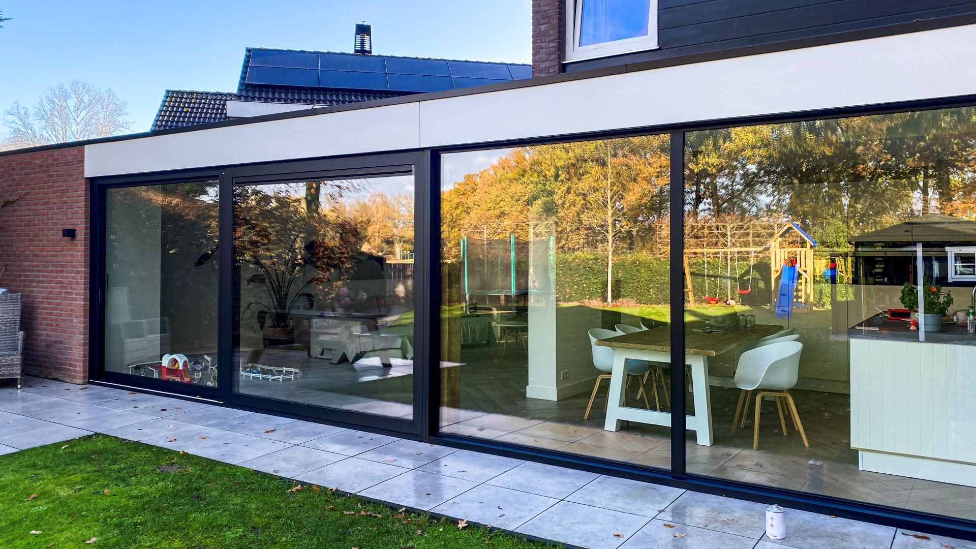 Brede aluminium schuifpui over de volledige gevel met uitzicht op een groene tuin en speeltoestellen