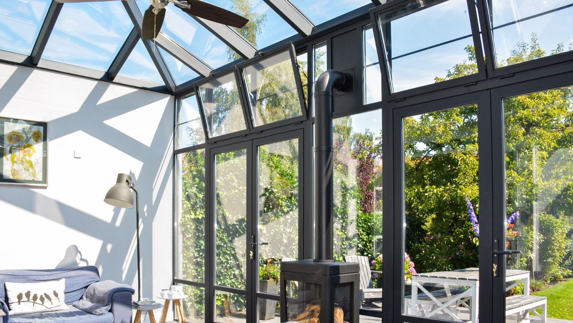 Lichte steellook serre met houtkachel, zwart aluminium kozijnen en uitzicht op groene tuin.