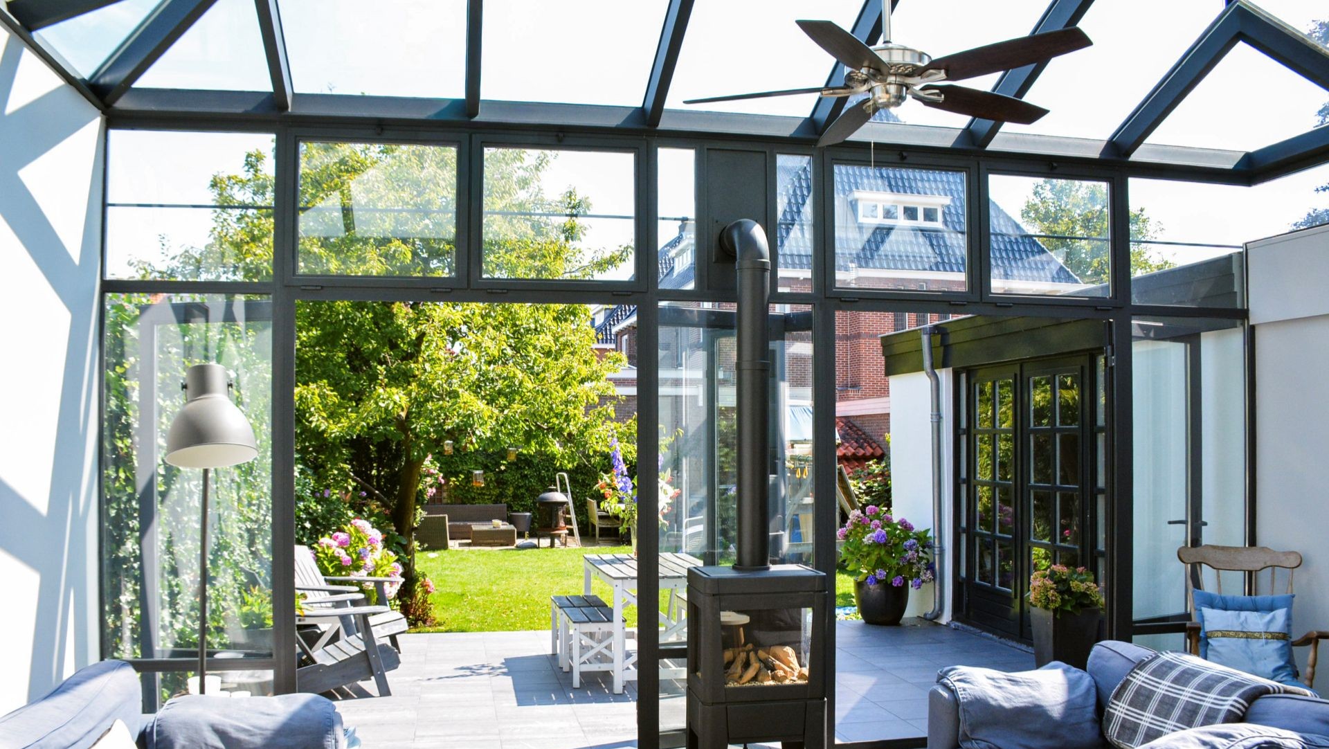 Interieur van steellook serre met glazen dak, houtkachel en open verbinding met de tuin.