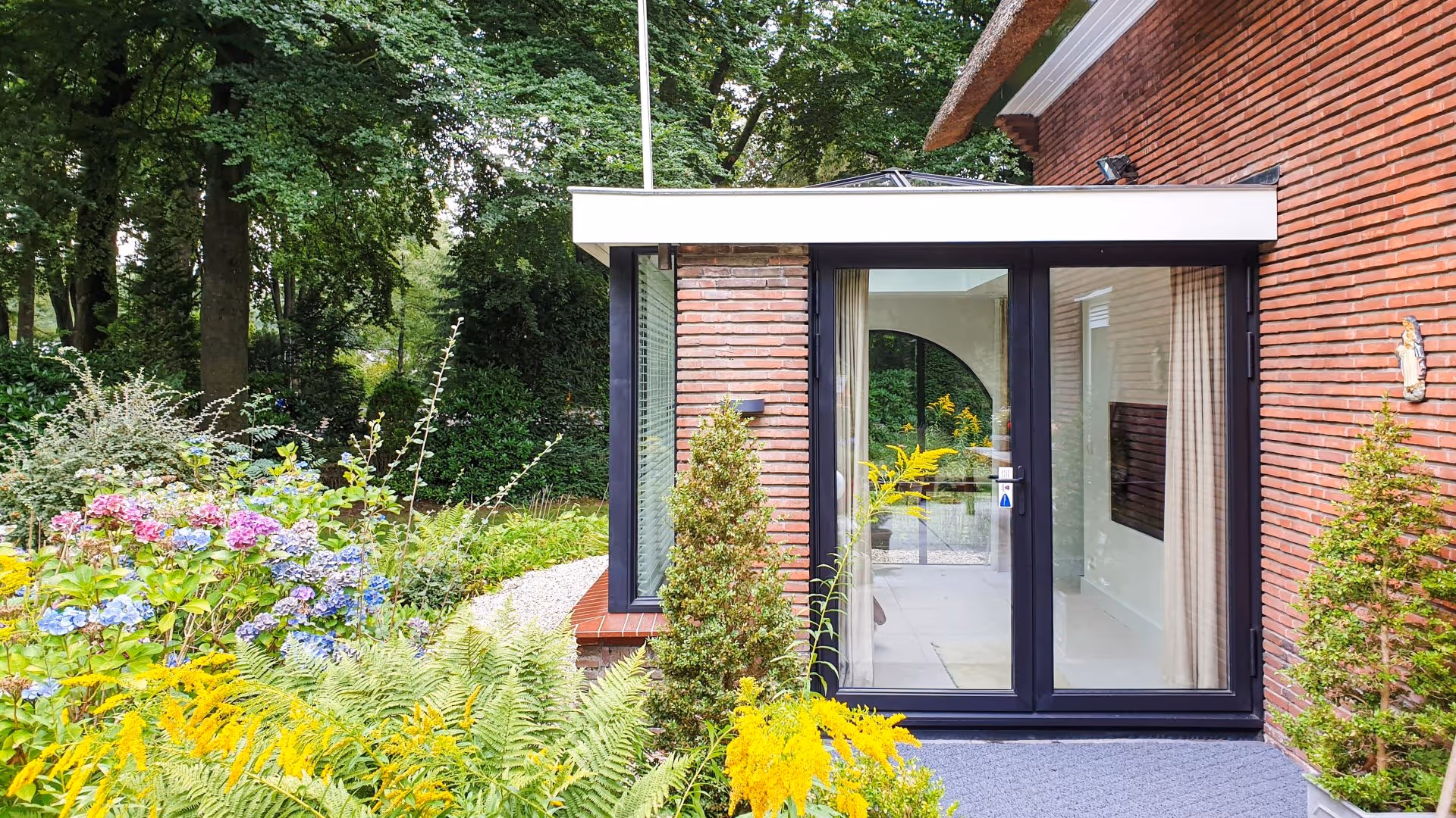 Moderne aanbouw met zwarte aluminium kozijnen, grote glaspartijen en uitzicht op groene tuin.