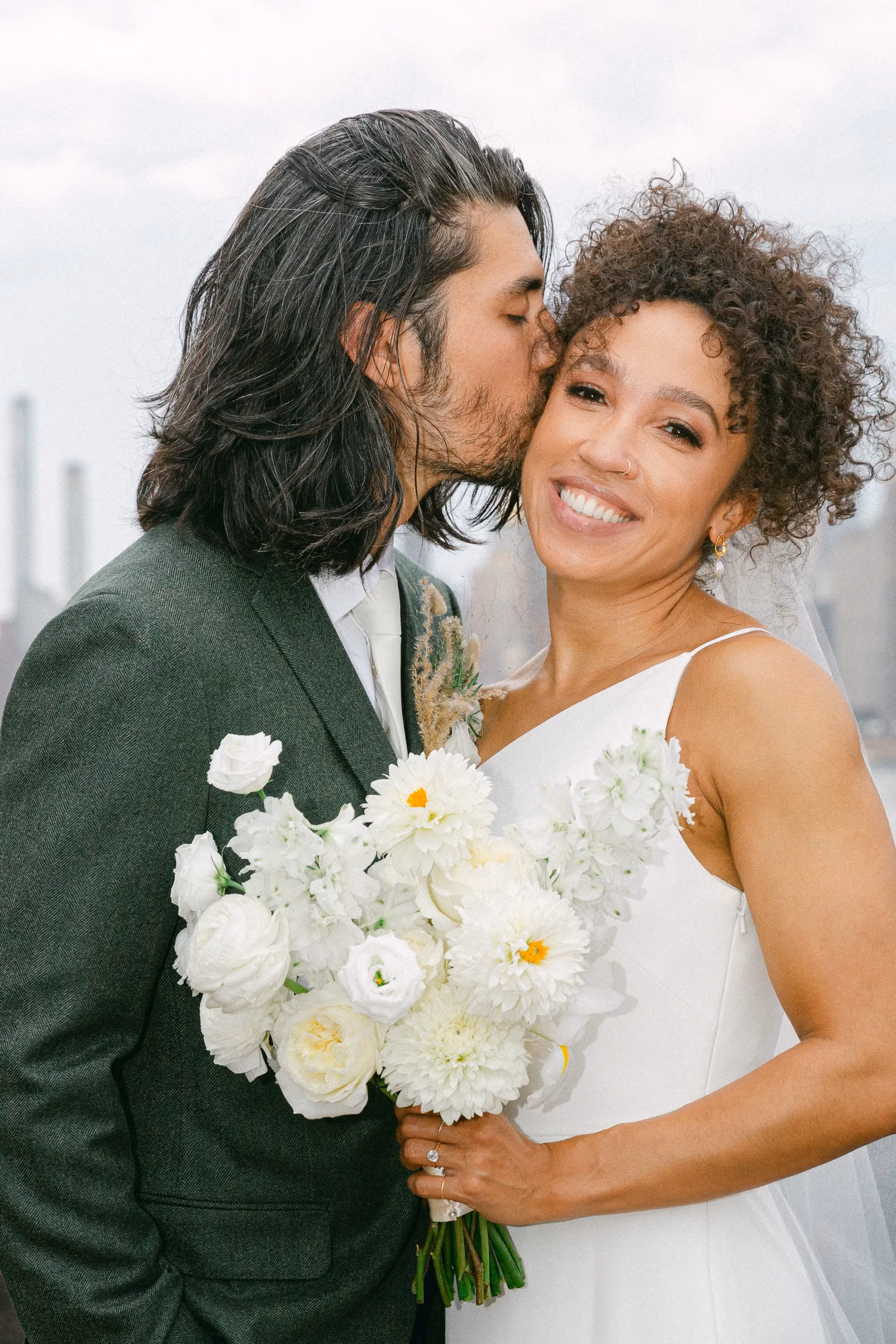 Groom kissing the bride's cheek in a rooftop portrait with the city skyline behind them.