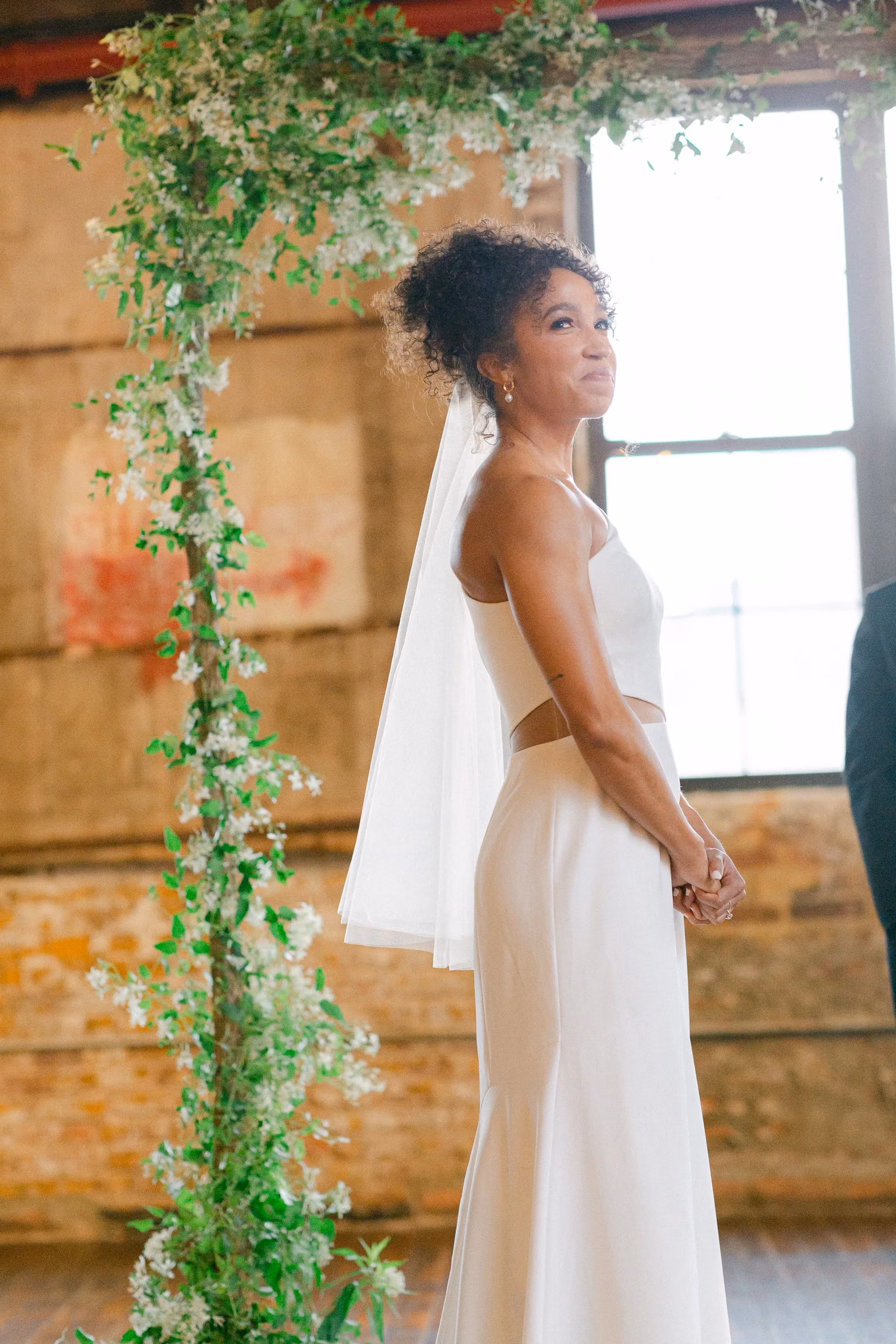 Bride glancing back with a smile beside the greenery-draped ceremony arch.