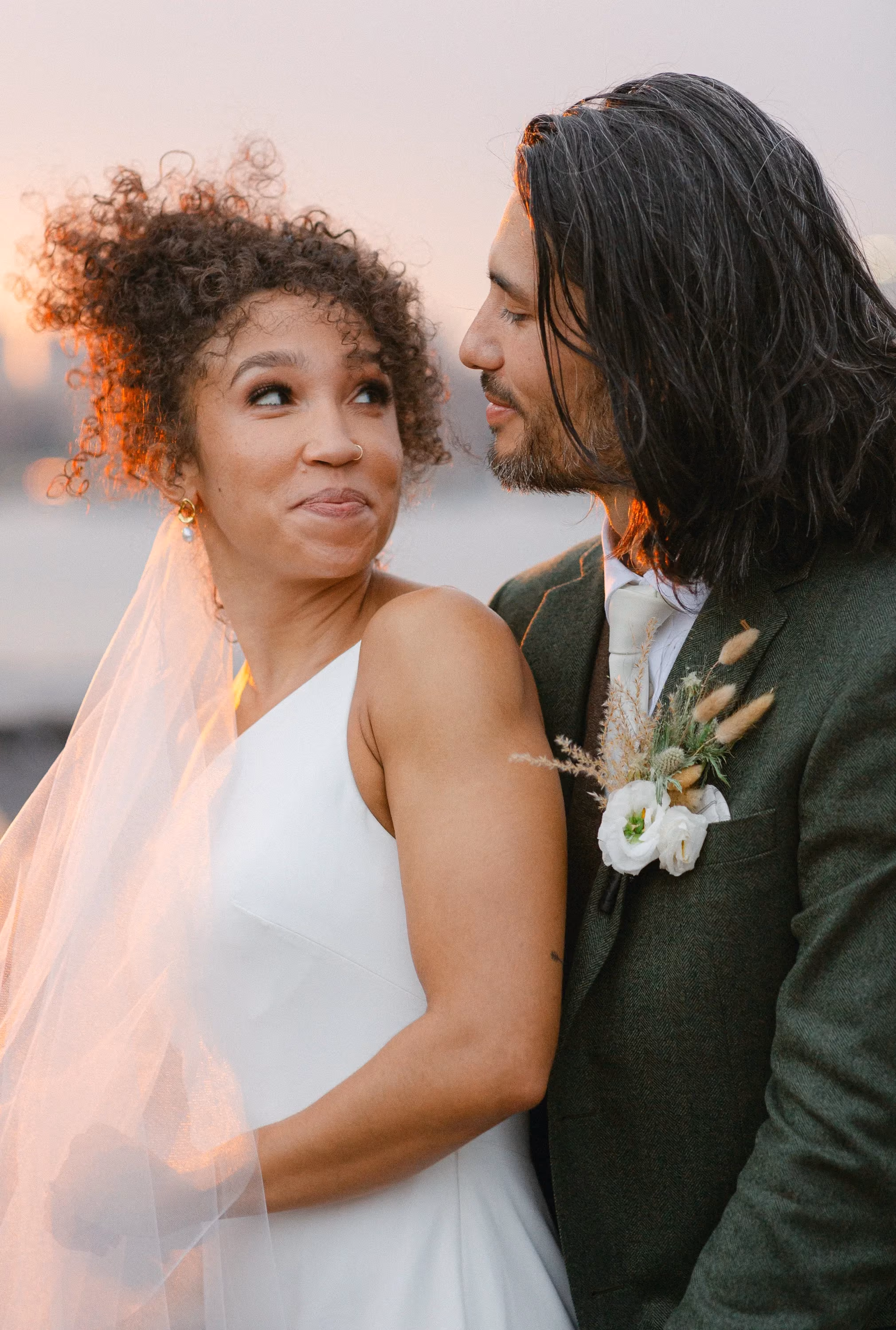 Bride and groom sharing a close embrace at sunset, her veil lifting in the breeze.
