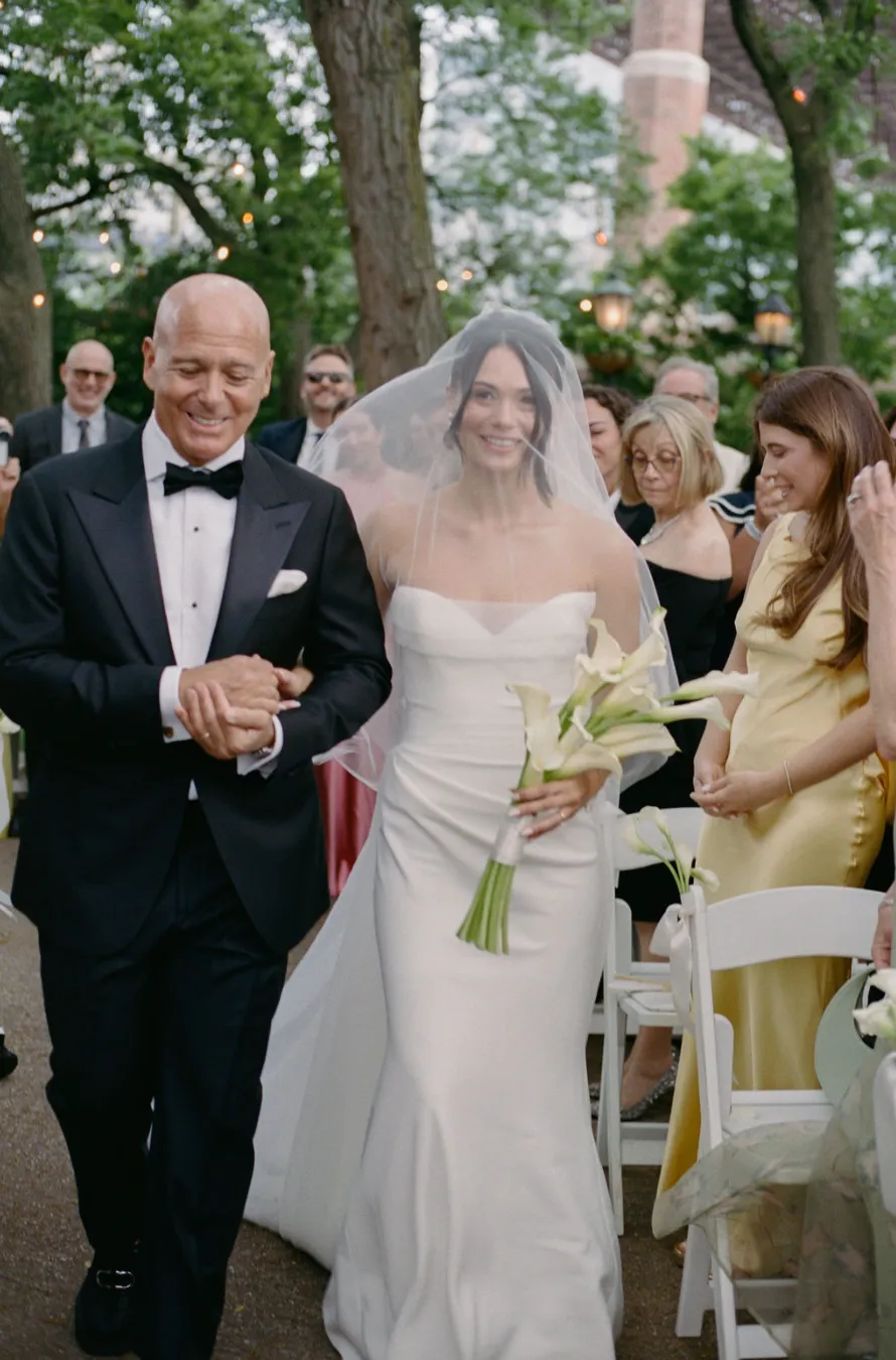 Bride walking down the aisle with her father, carrying a calla lily bouquet, guests seated nearby