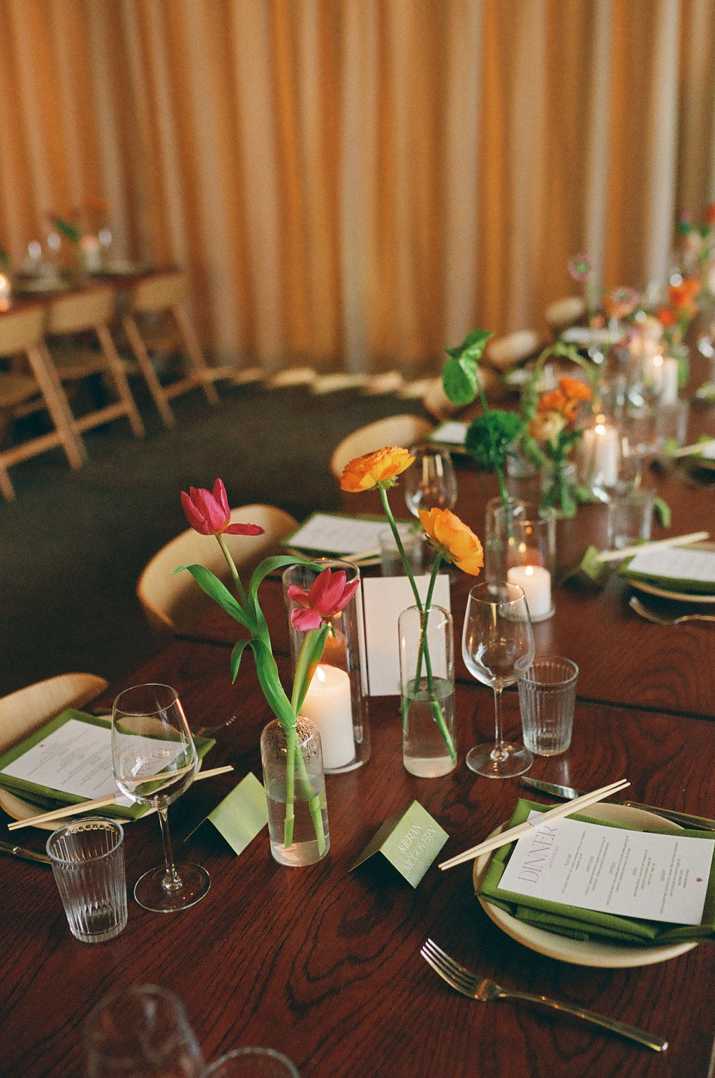 Long reception table with bud vases of red tulips and orange ranunculus among candlelight