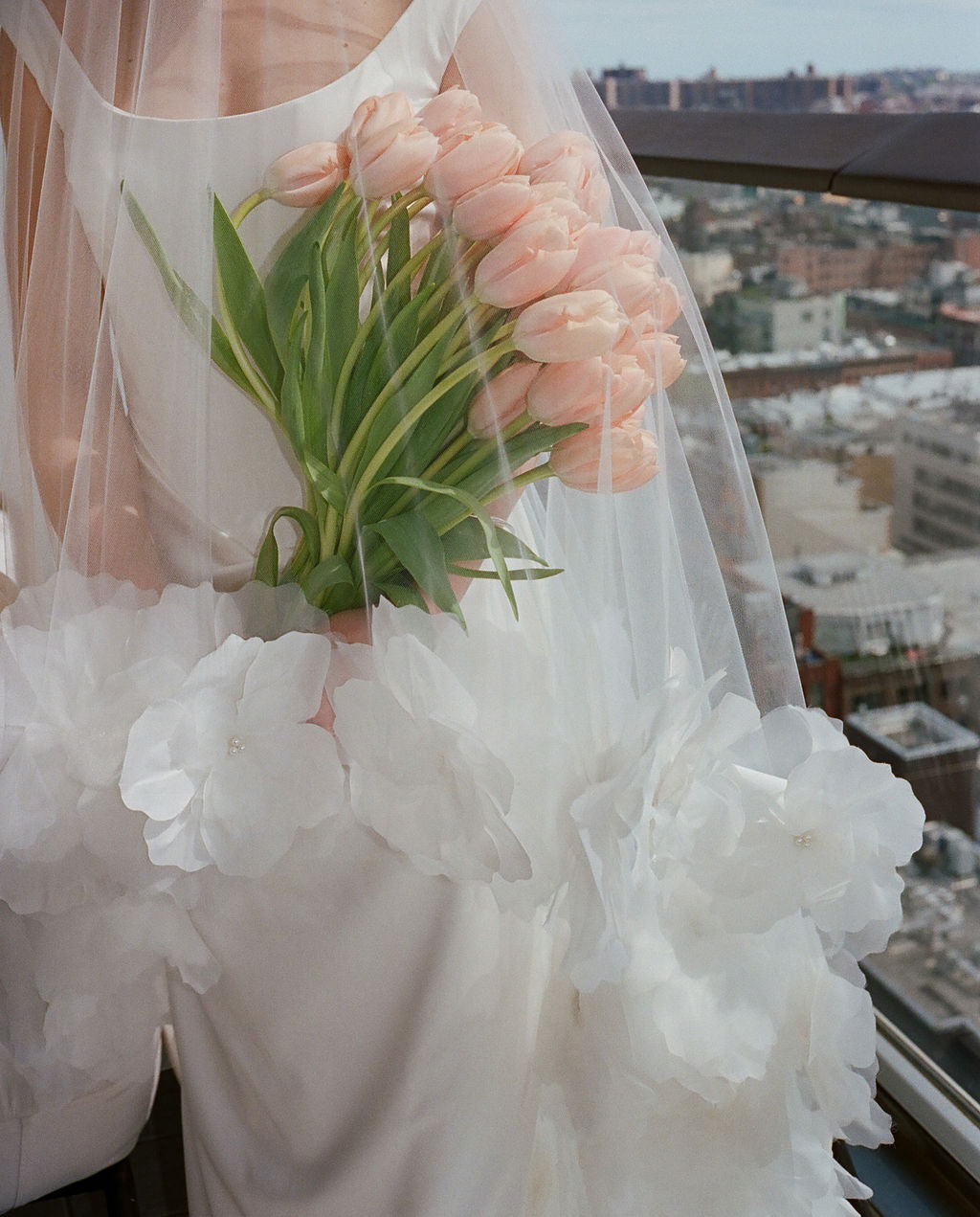 Bride holding a bouquet of pink tulips against her floral-appliqué gown with the city skyline in the background