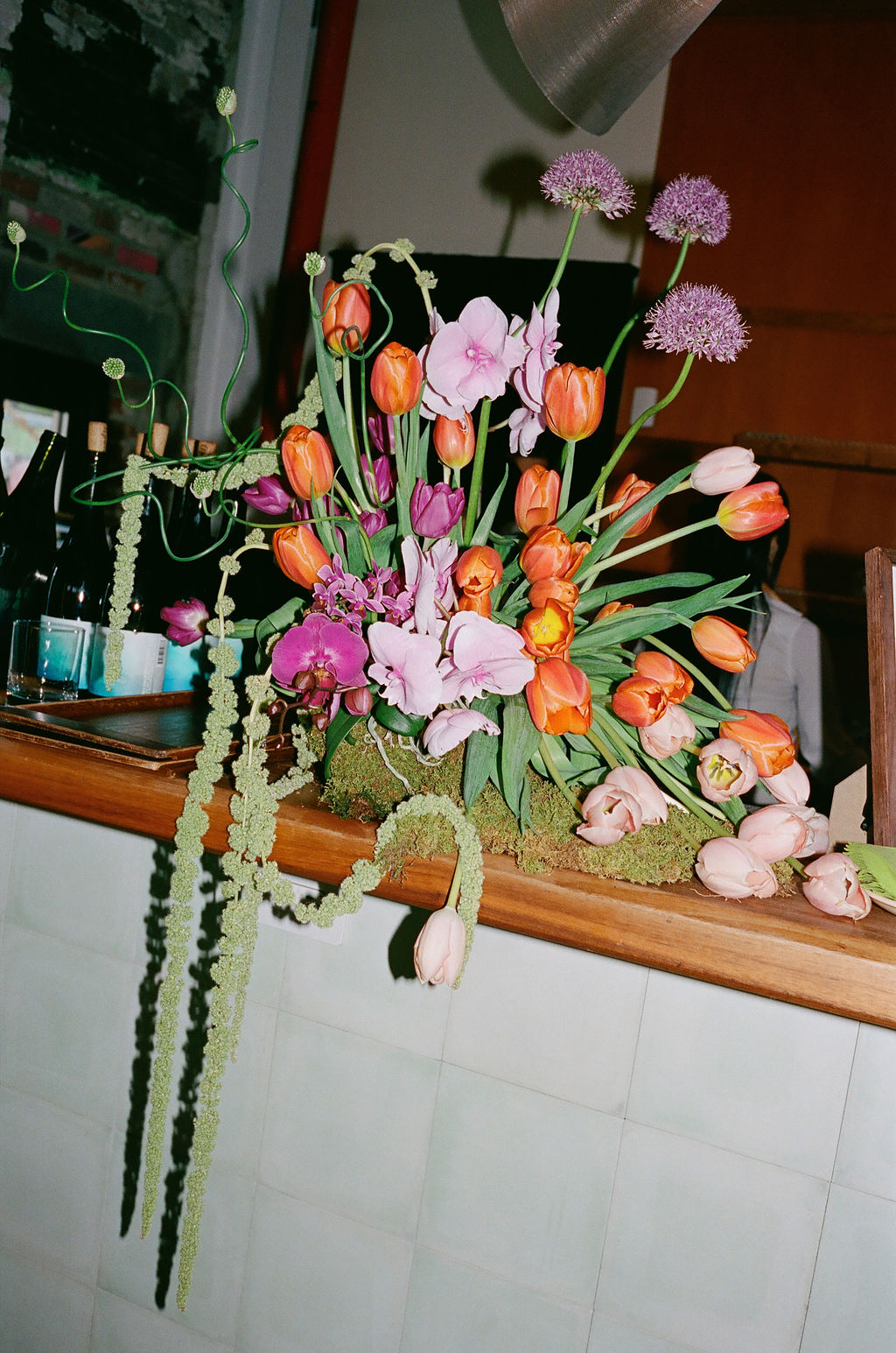 Bar-top floral arrangement with orange tulips, purple allium, orchids, and trailing amaranthus