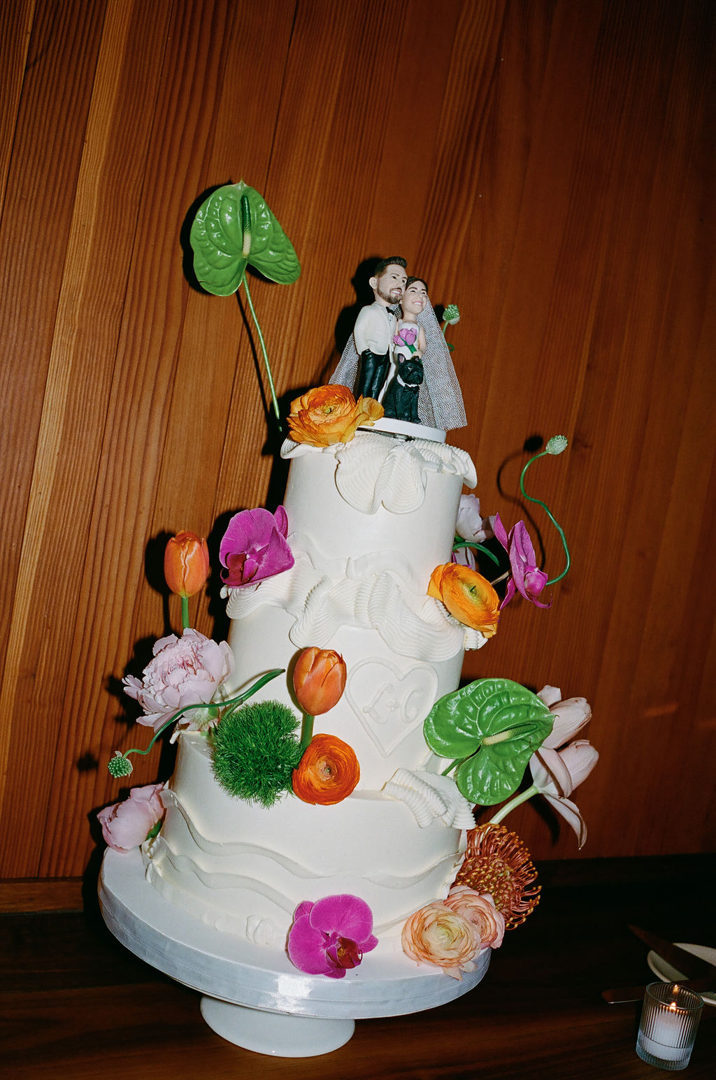 White tiered wedding cake with a bride and groom topper, decorated with tulips, ranunculus, and orchids