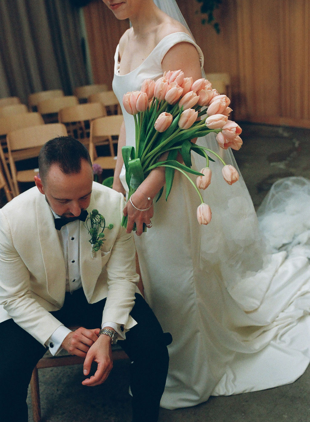 Bride holding a bouquet of pink tulips beside her seated groom in a cream tuxedo