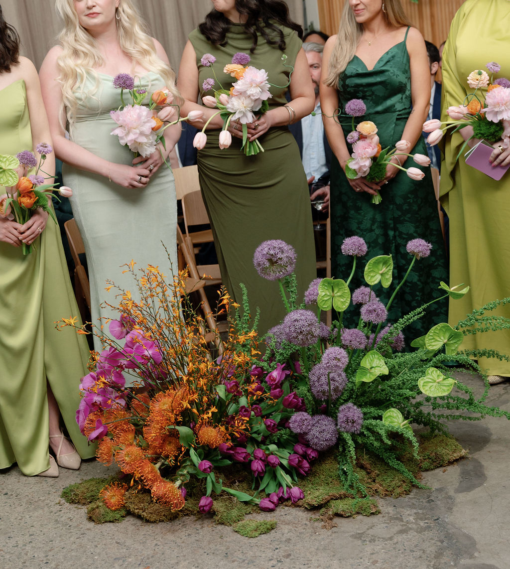 Bridesmaids in green dresses holding bouquets, standing beside a colorful ceremony floor arrangement