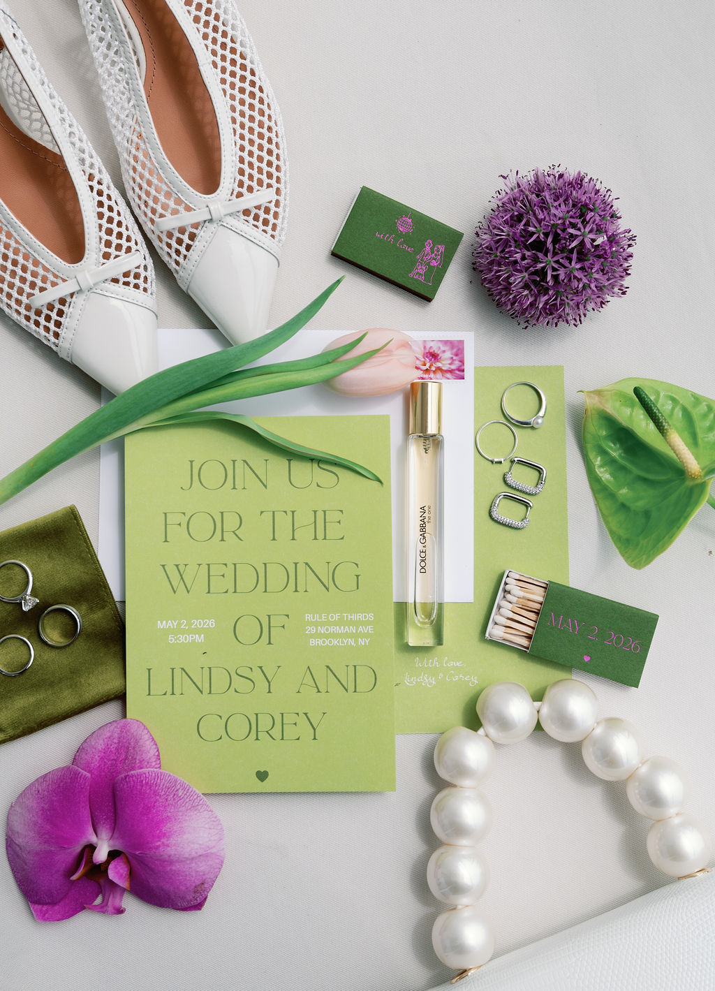 Flat lay of a wedding invitation suite styled with purple allium, orchid, and pearl jewelry