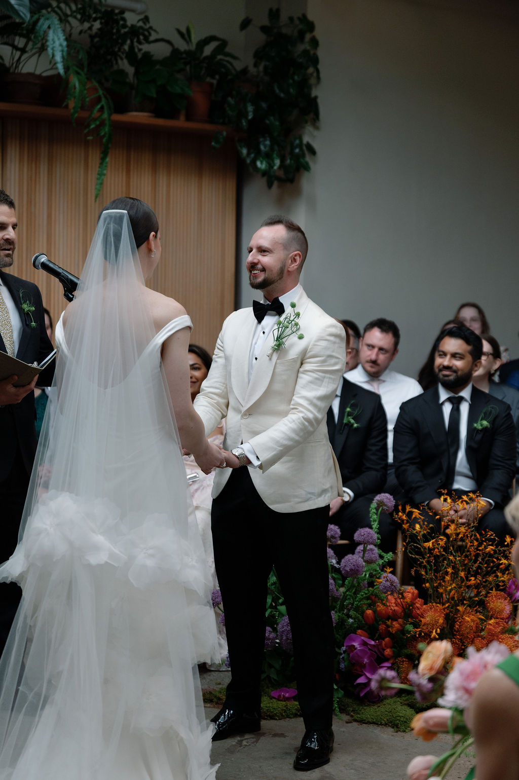 Groom smiling at his bride during the ceremony vows, officiant and guests looking on