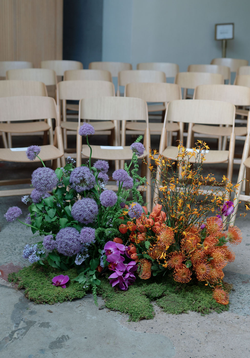 Ceremony floor arrangement of purple allium, orange pincushion protea, and orchids beside rows of wooden chairs