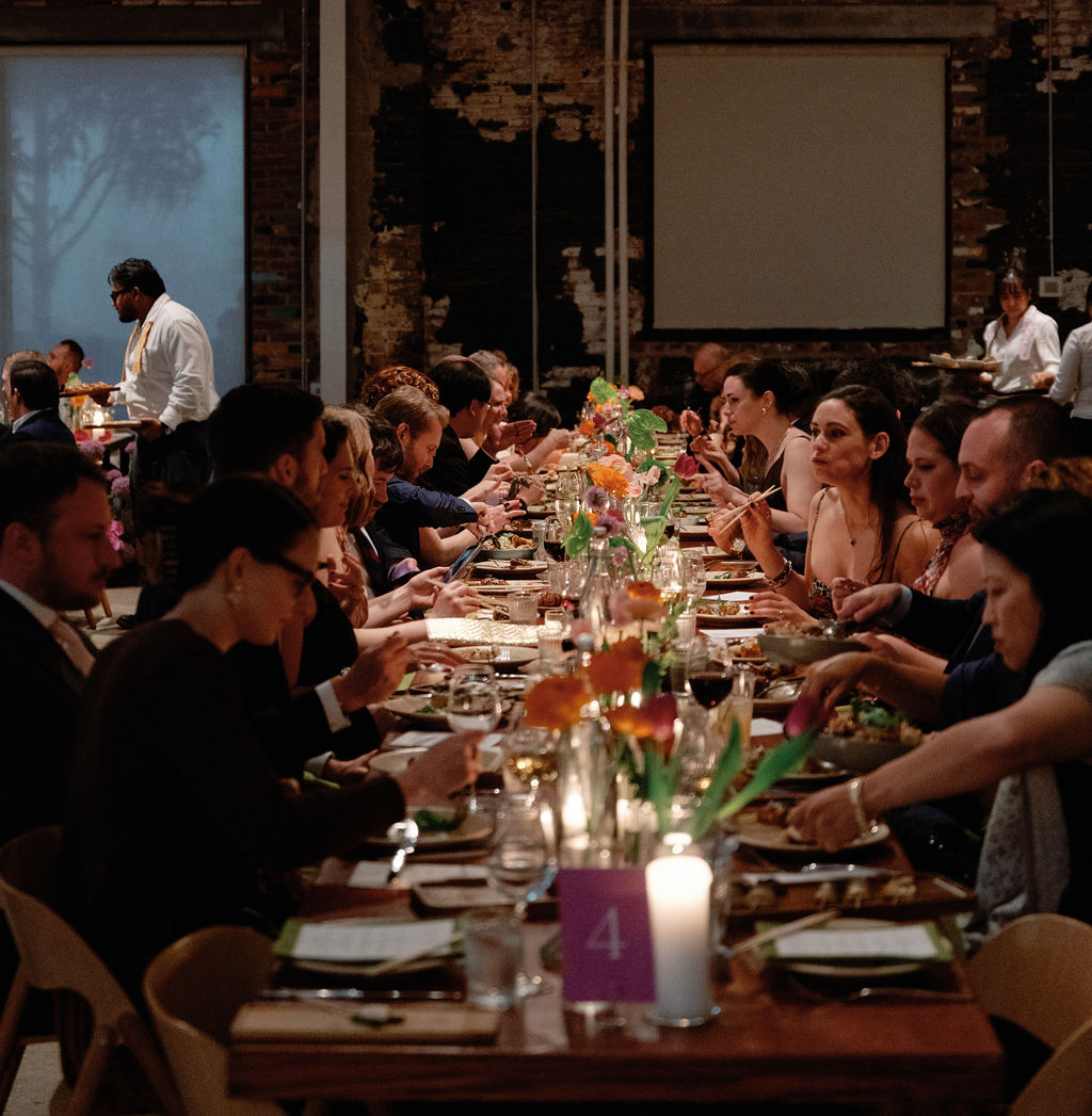 Guests dining at the long reception table during dinner service, exposed brick wall behind