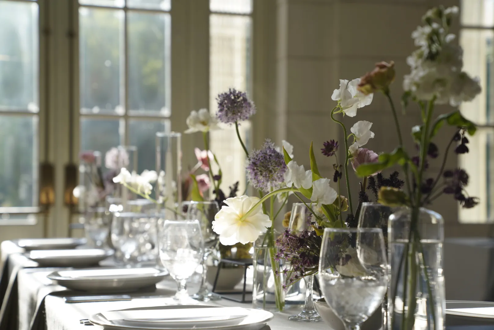 Reception tablescape with bud vases of white sweet peas, purple allium, and garden roses near the windows.