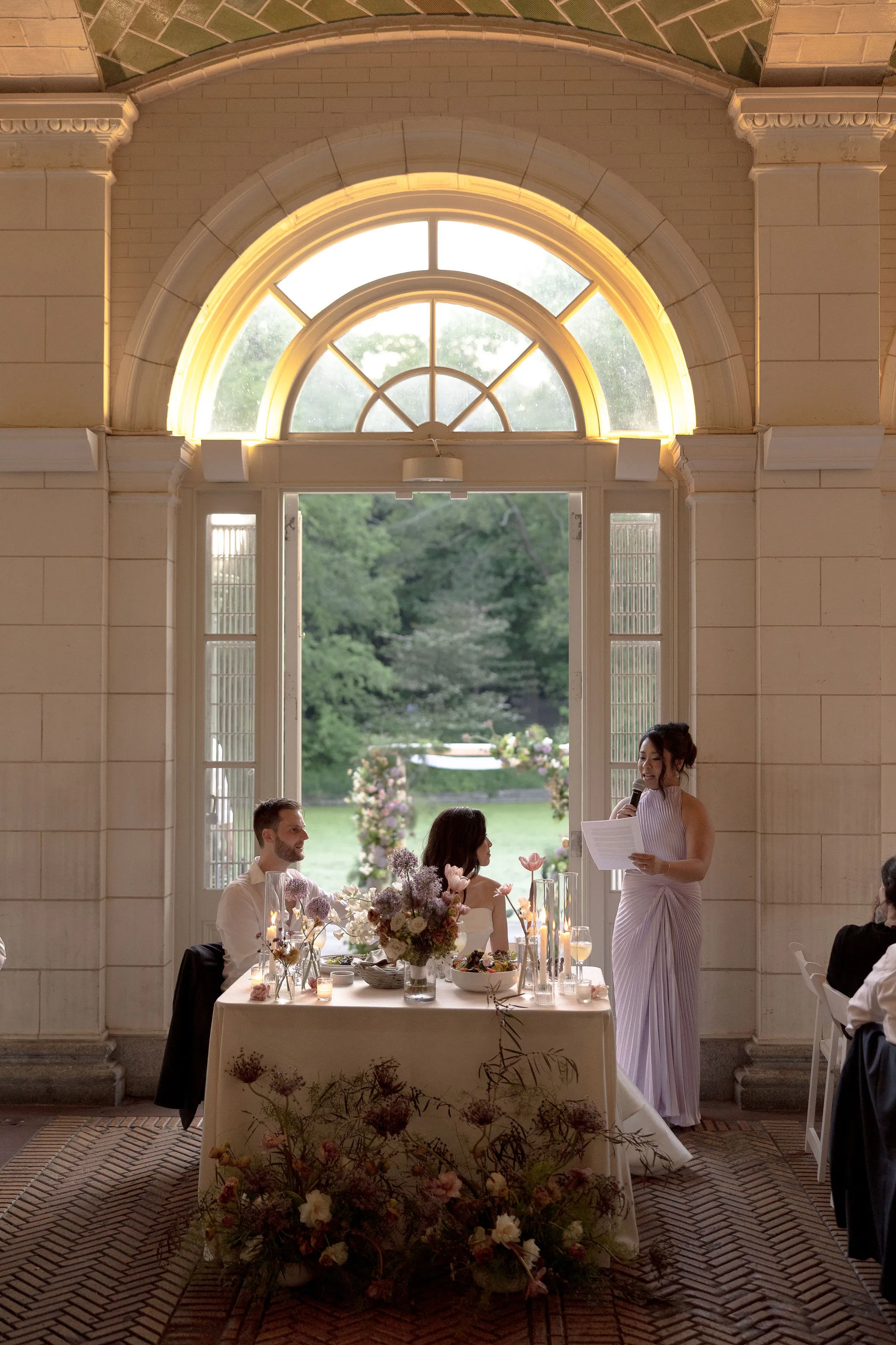 Groom gives a toast at the sweetheart table, with the ceremony arch and lake visible through the open doorway.