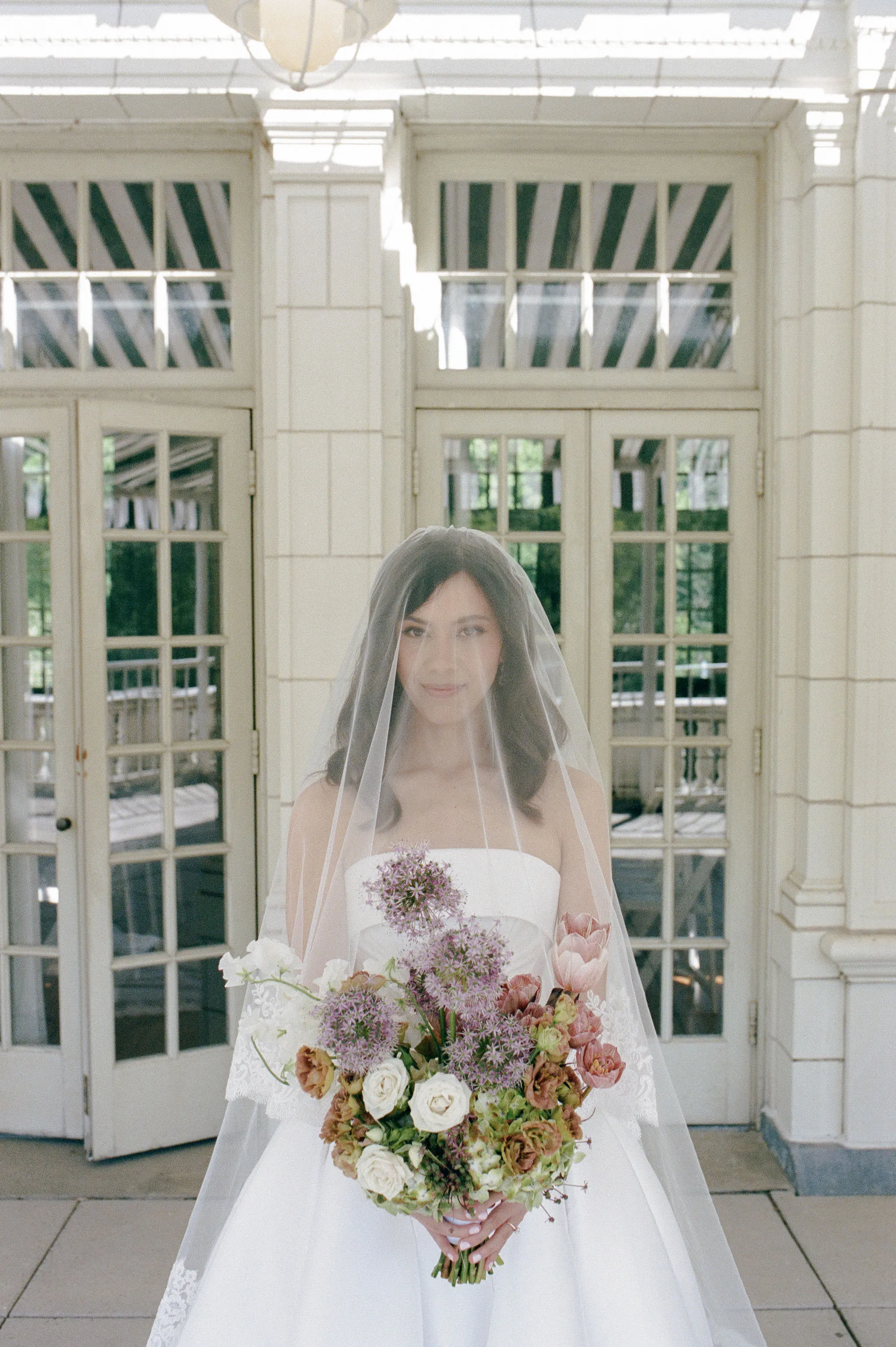 Bride in a cathedral veil holding an allium and garden rose bouquet in front of the Boathouse's arched glass doors.