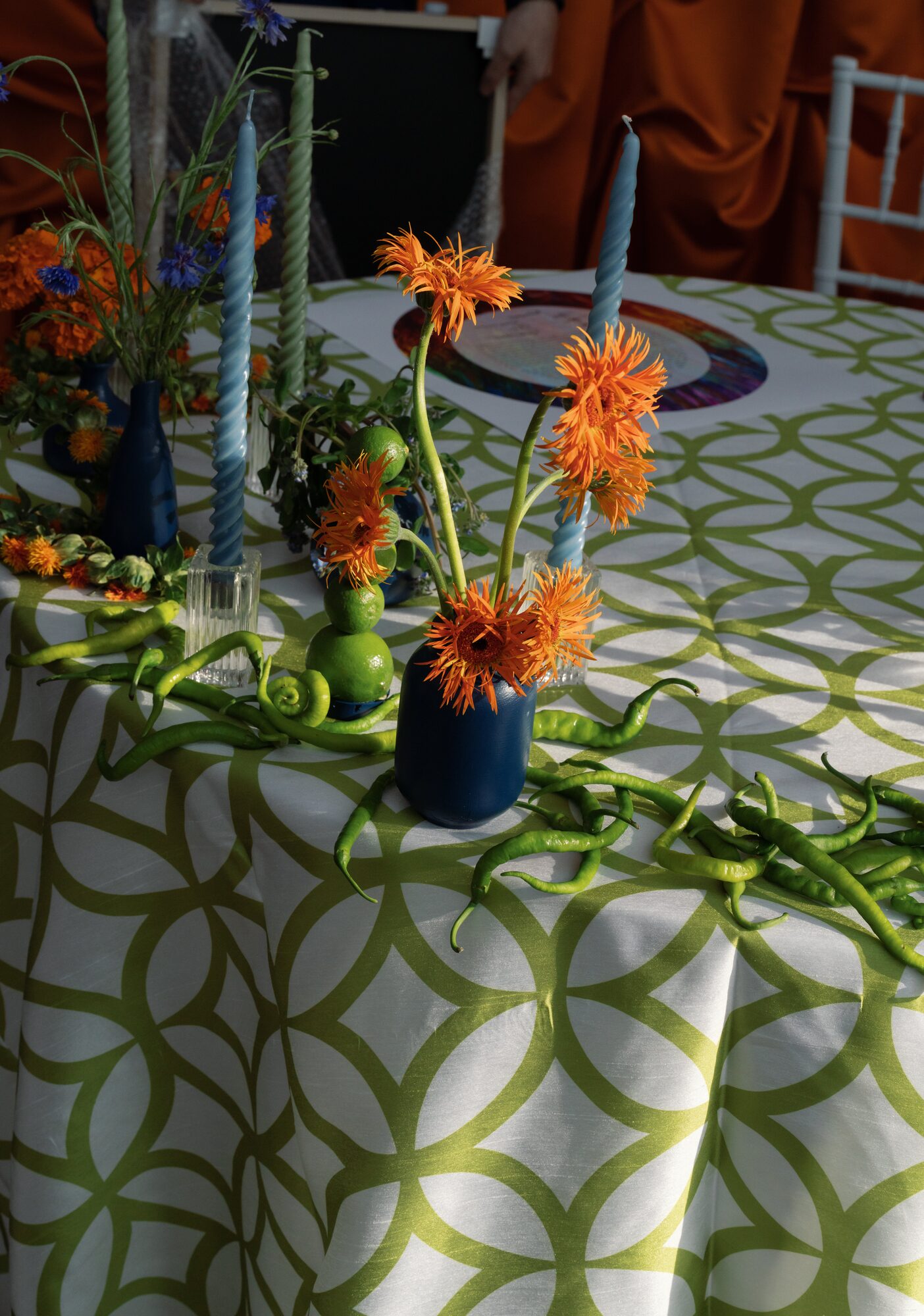 Bold orange, green and blue reception decor with gerbera daisies in navy vases, blue taper candles and trailing fresh green chile peppers on a quatrefoil linen.