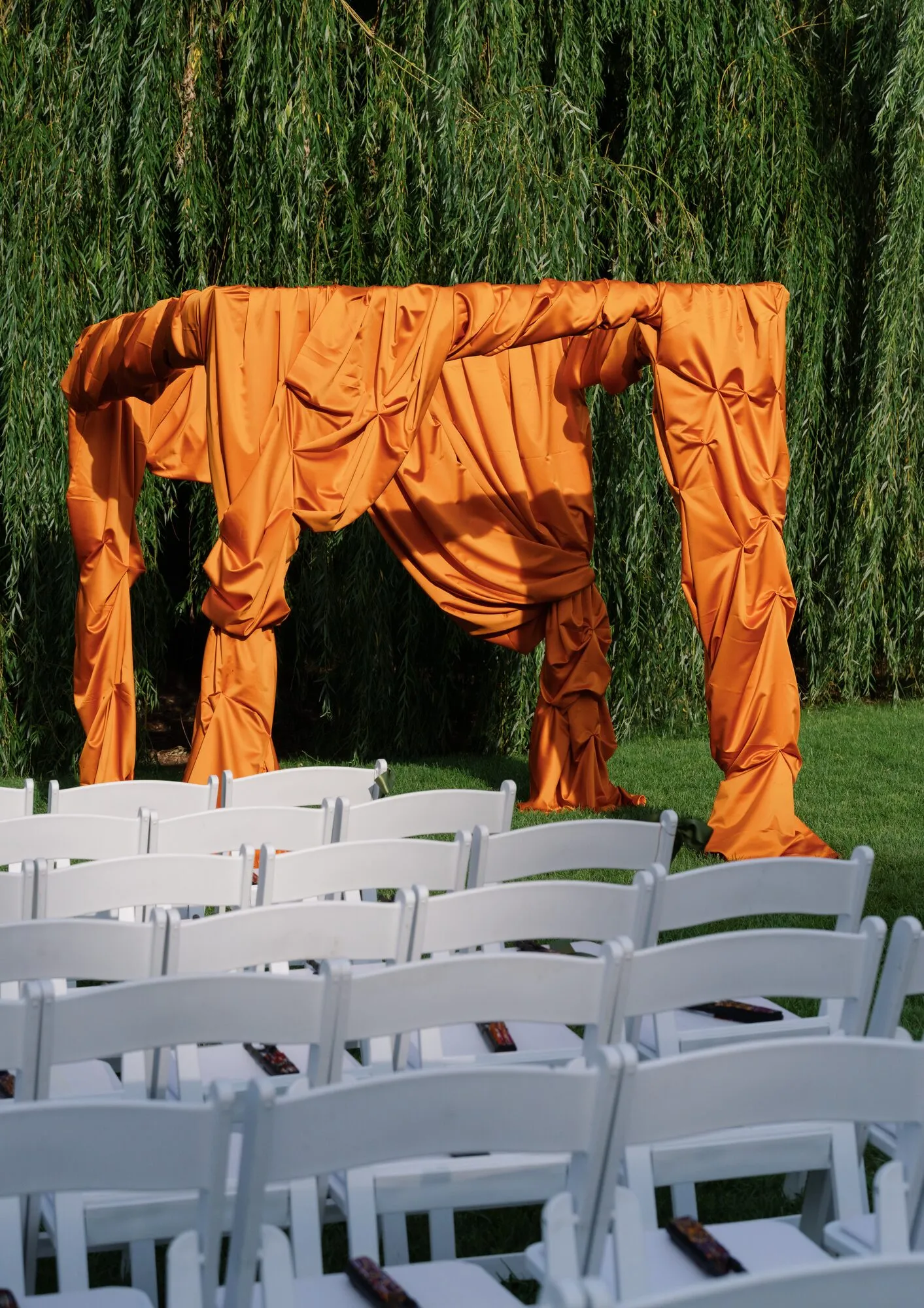 Chuppah draped in bold orange fabric set against a weeping willow tree, flanked by rows of white folding chairs.
