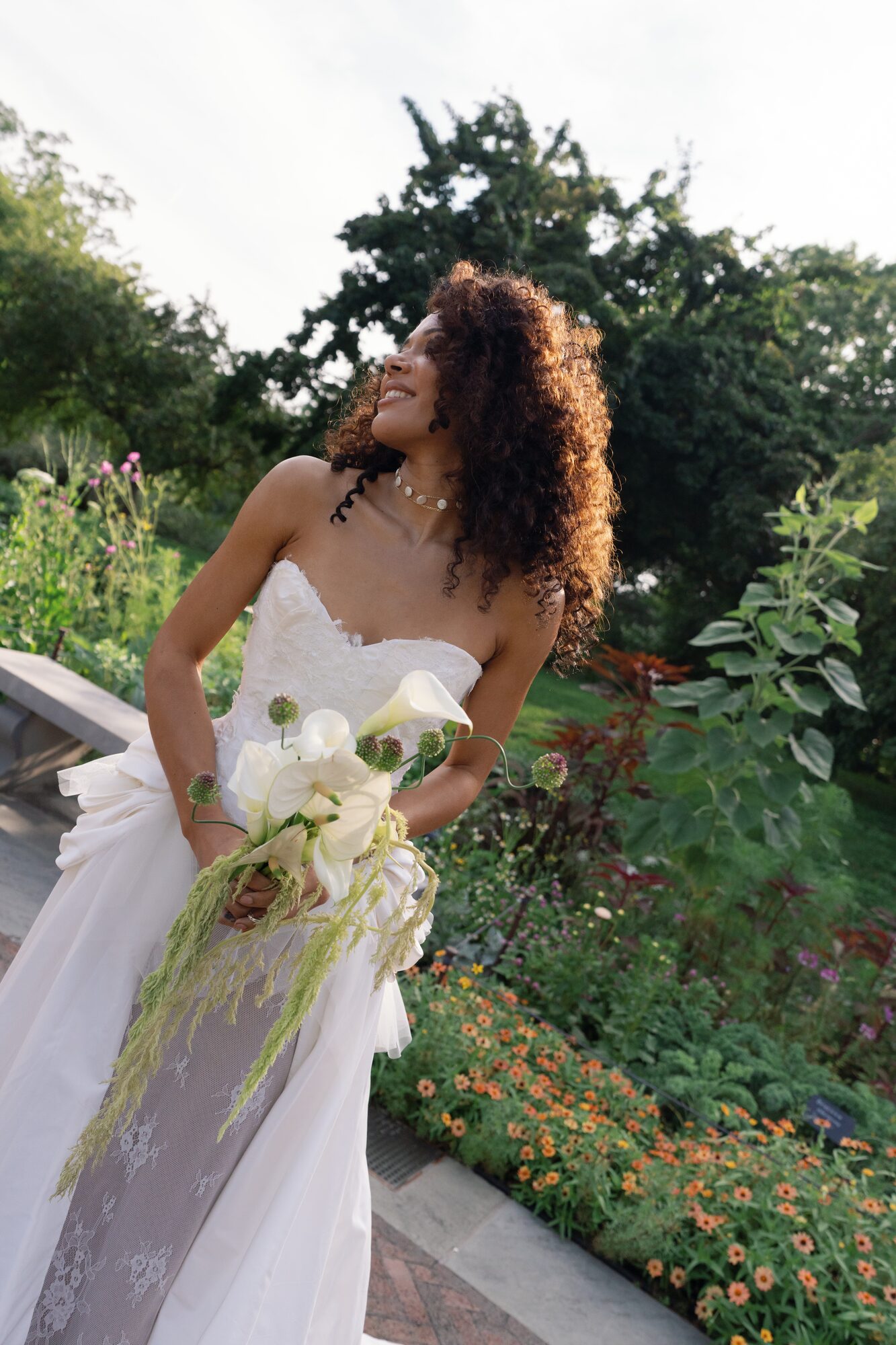 Full-length garden portrait of the bride holding a textured bridal bouquet of calla lilies, allium and hanging amaranthus, with orange garden blooms behind her.