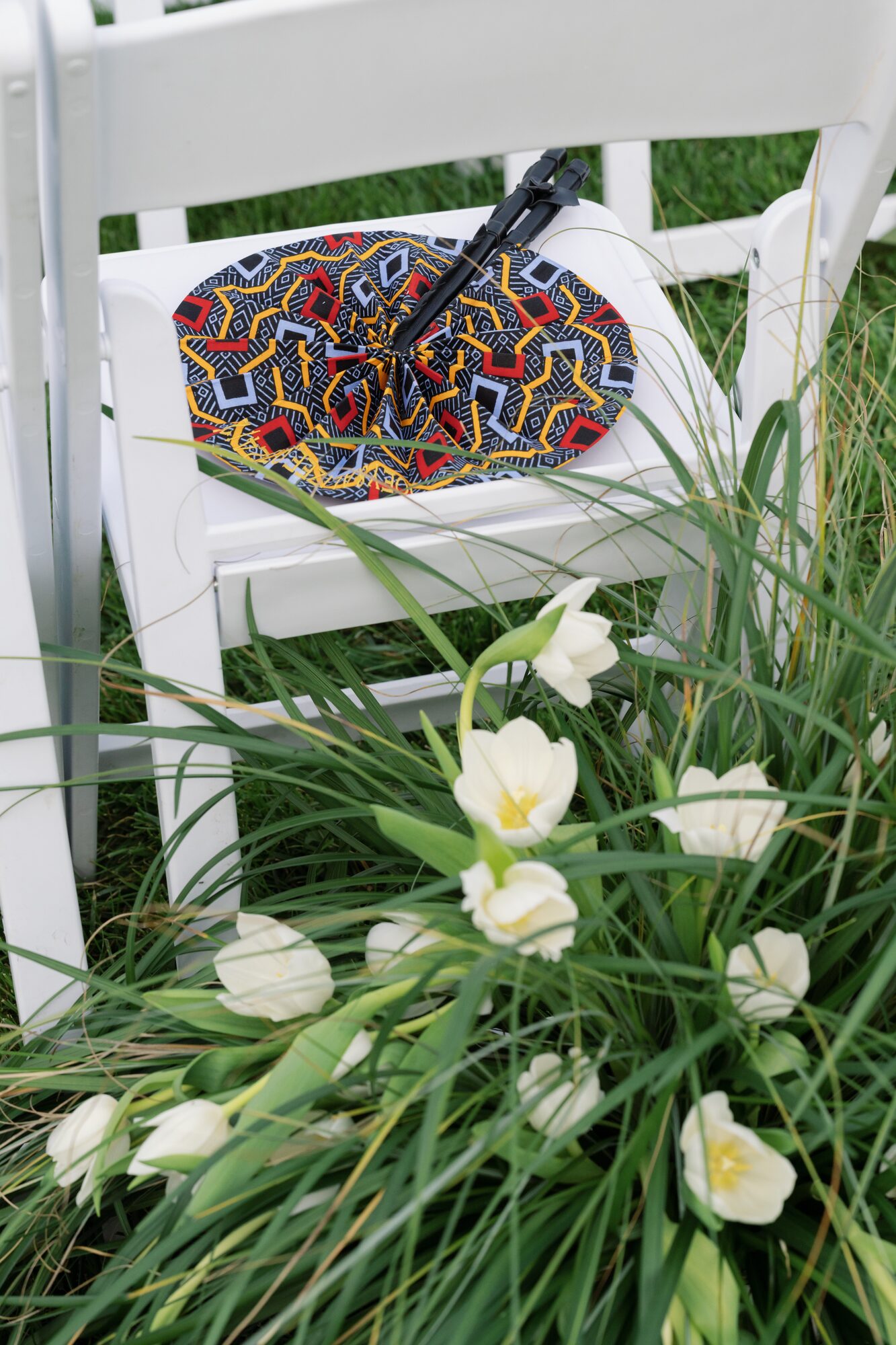 Detail shot of a white folding ceremony chair with a bold graphic-print fan, surrounded by white tulips and ornamental grass.