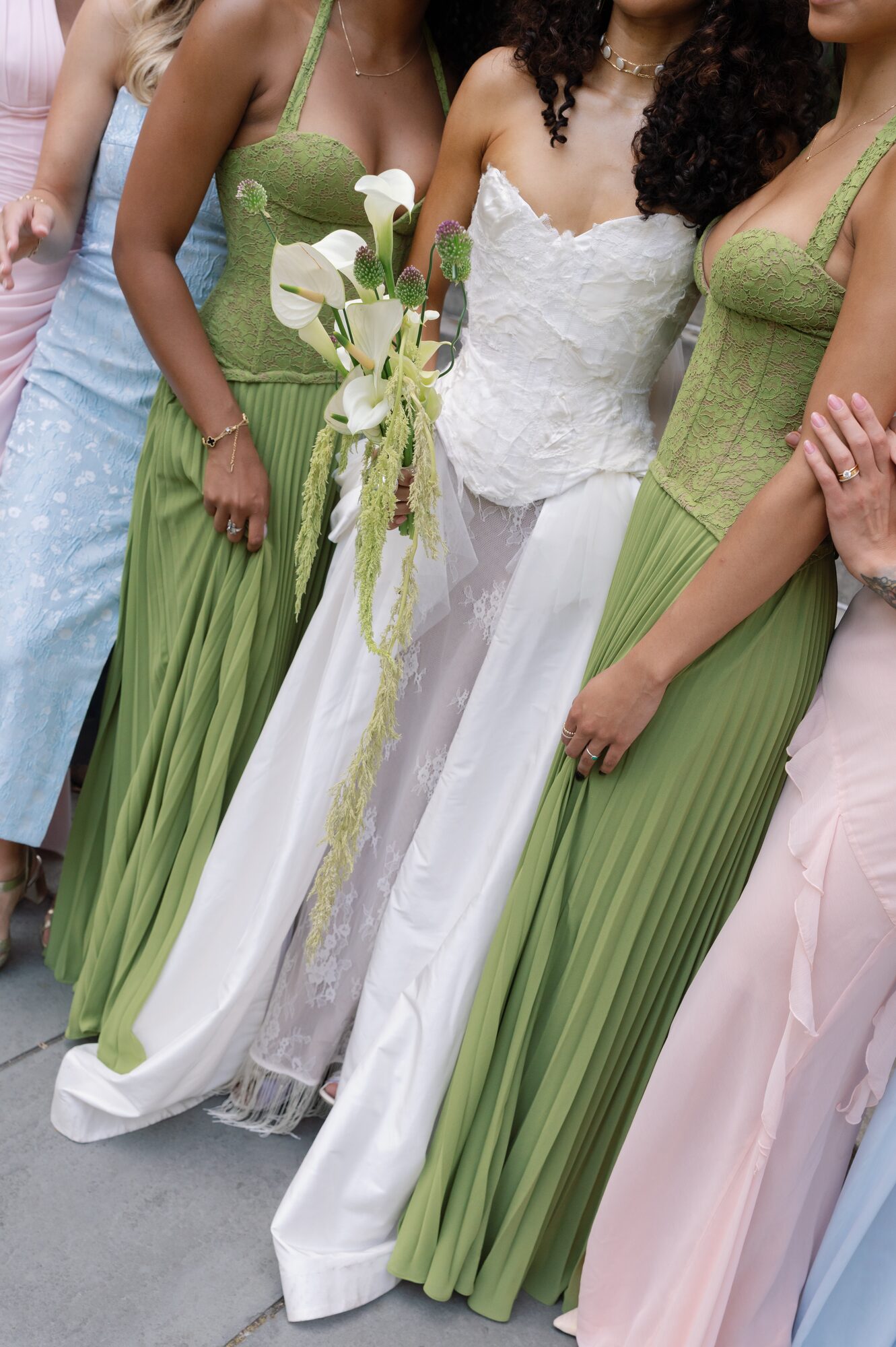 Bride in a strapless lace gown standing between bridesmaids in sage green dresses, holding her calla lily and allium bouquet with trailing amaranthus.