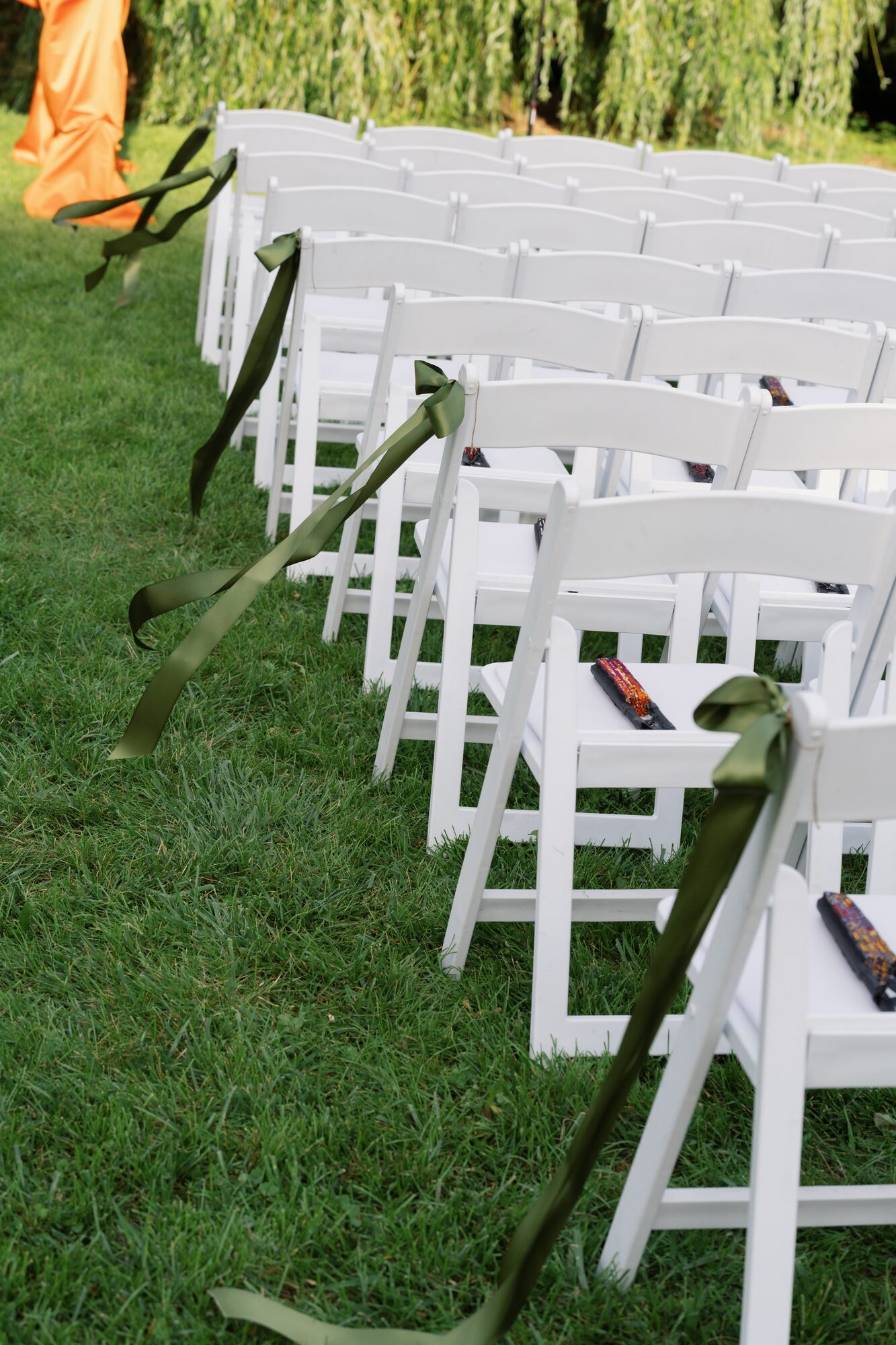 Row of white ceremony chairs tied with trailing olive green ribbon along the grass aisle.