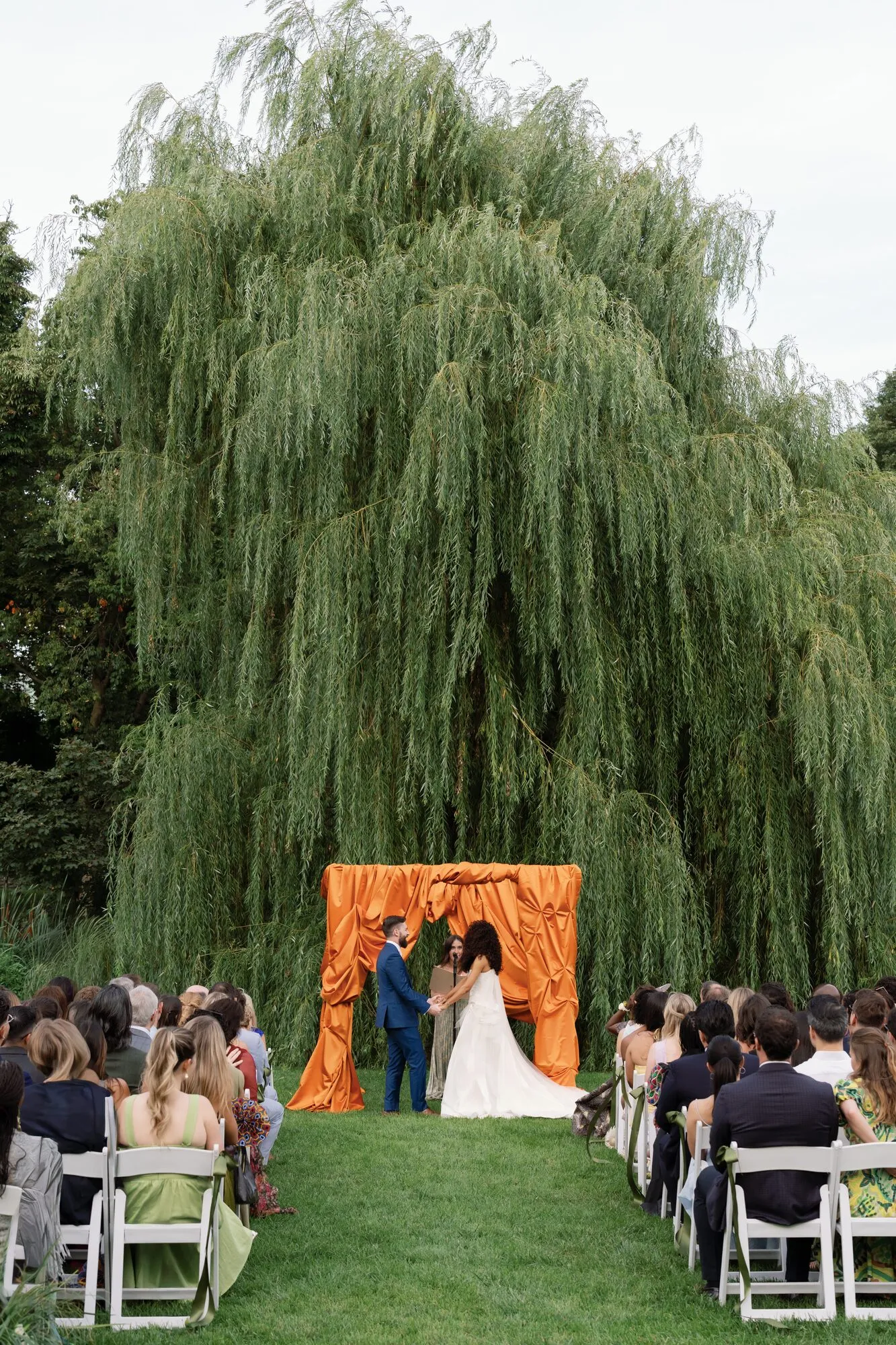 Wide ceremony view of the couple exchanging vows under an orange-draped chuppah beneath a willow tree, guests seated on white chairs.