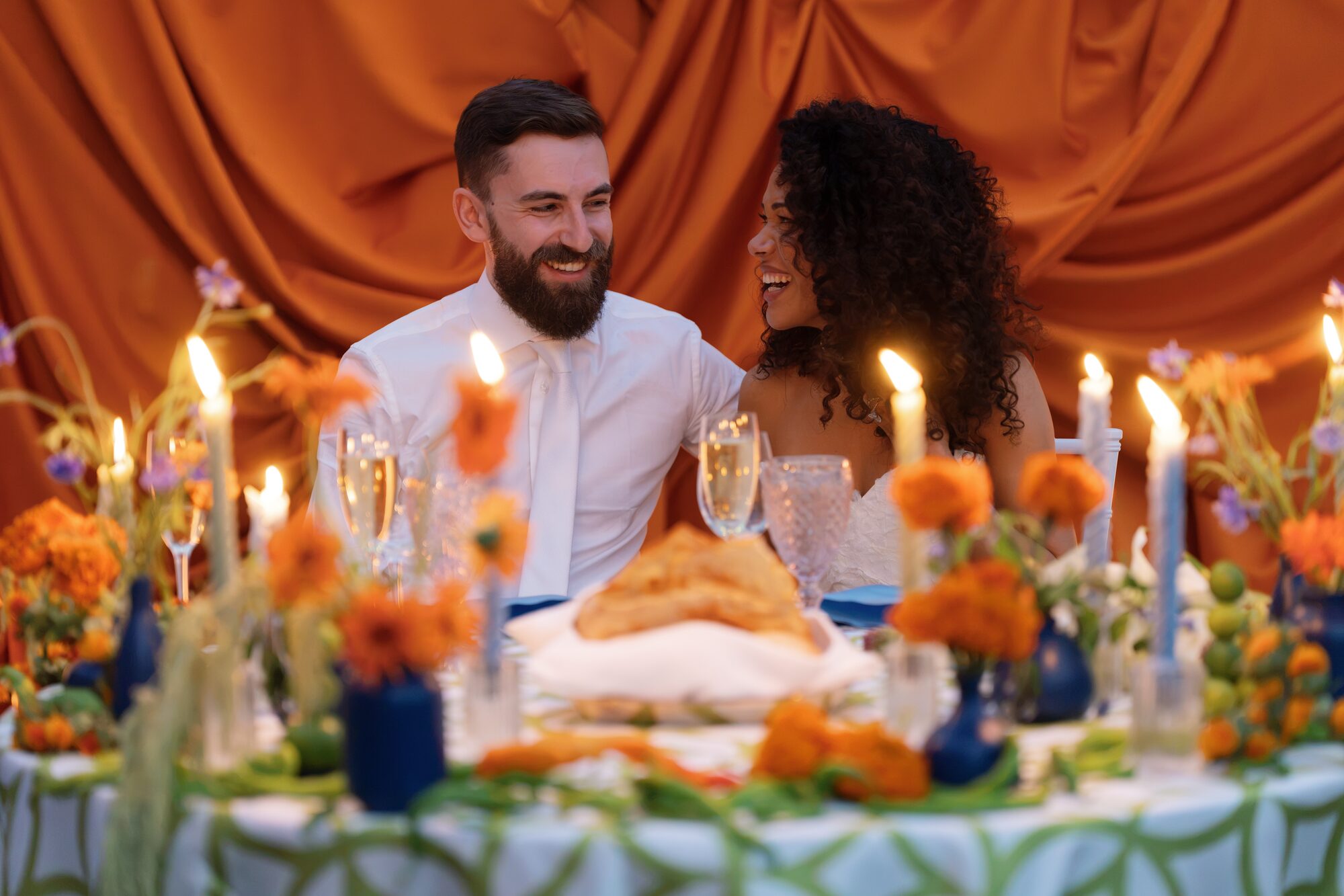 Bride and groom laughing together at their sweetheart table, framed by the orange chuppah backdrop and glowing taper candles.